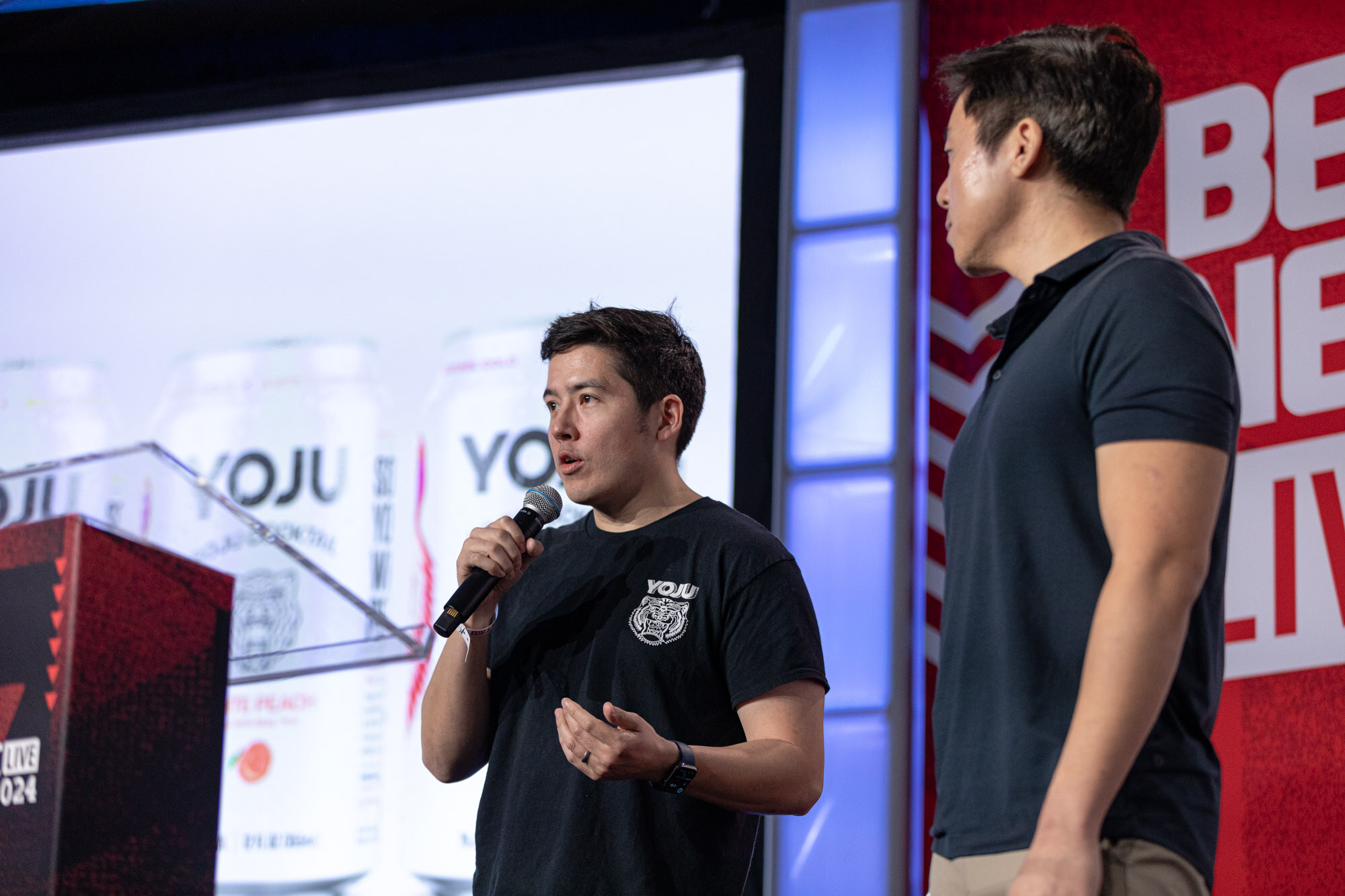 Man in a black Yoju t-shirt speaks into a microphone at a podium with a Yoju slide behind him.