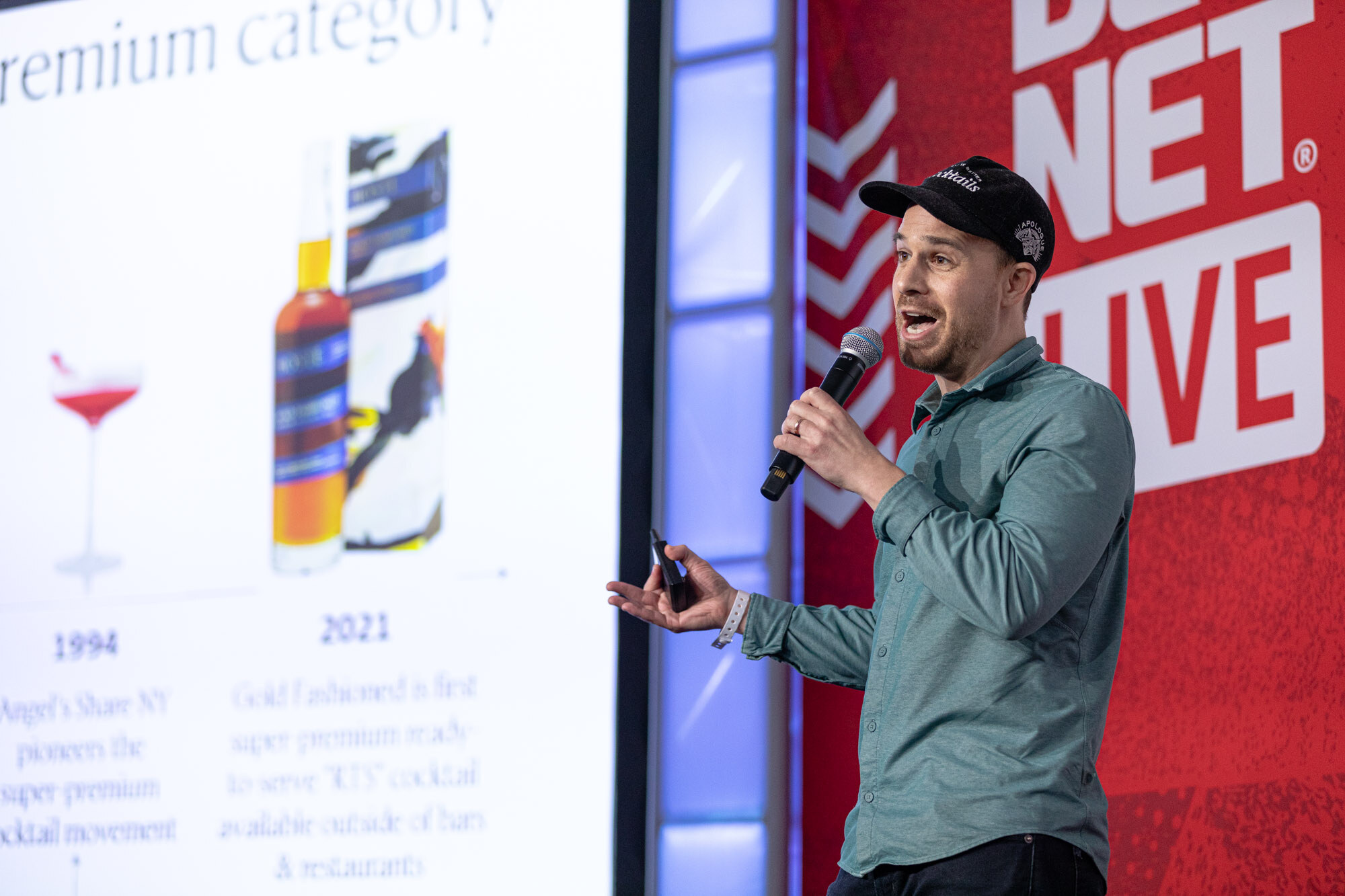 Man in a teal shirt and cap holds a microphone and gestures in front of a slide about premium ready-to-drink cocktails.