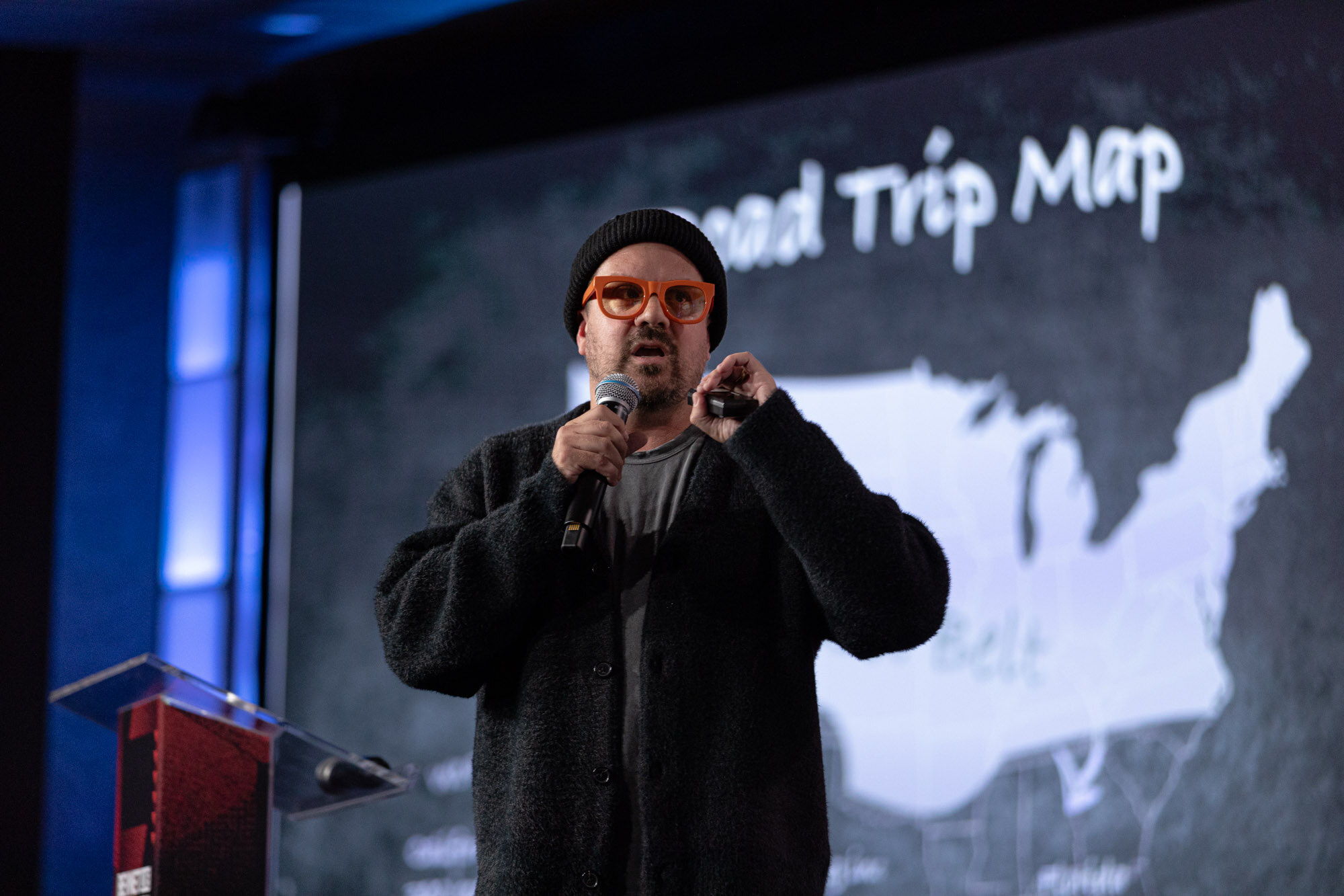Man in a black beanie and orange glasses speaks in front of a US map slide.