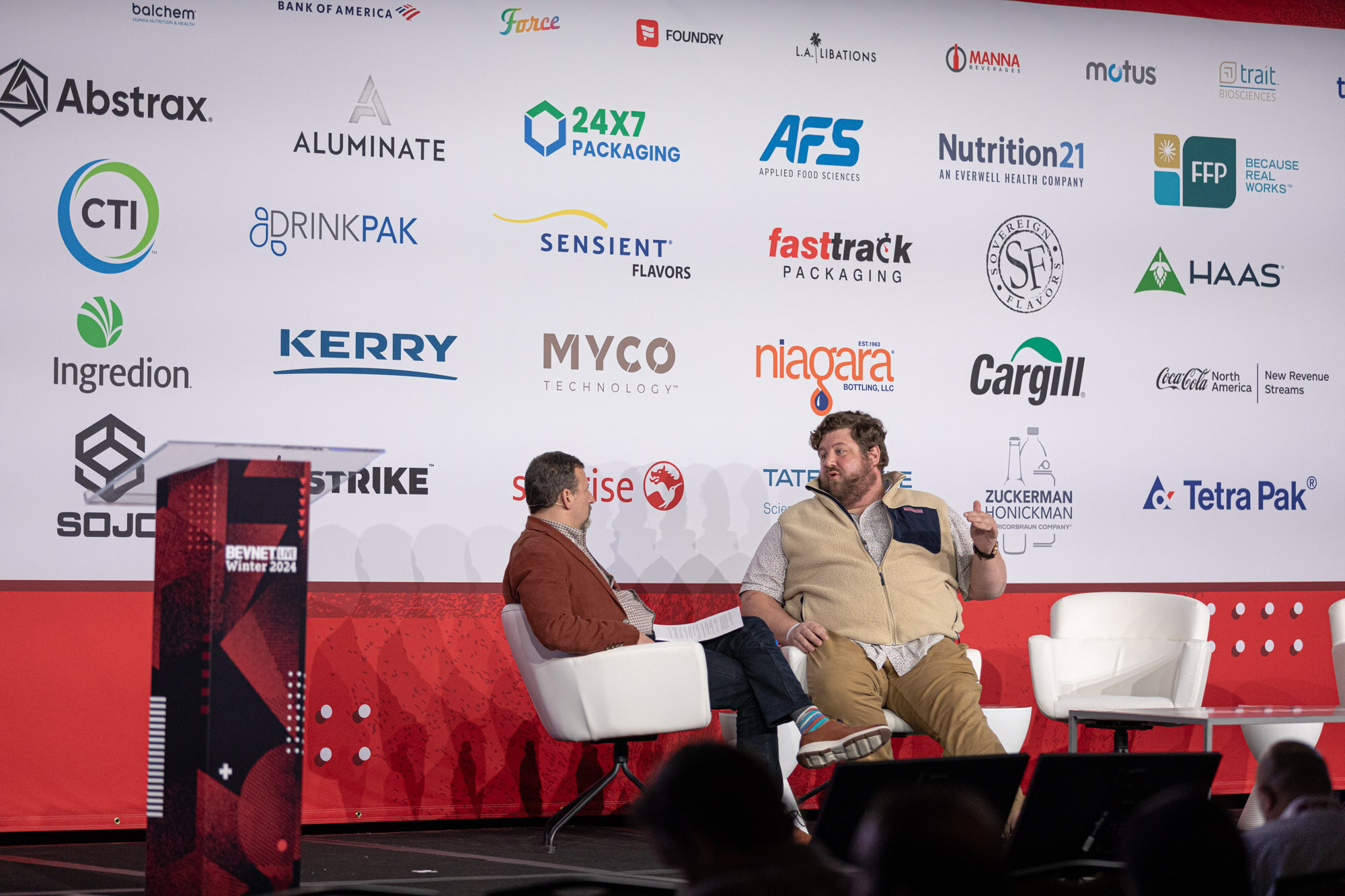 Two men sit in white armchairs on a stage with a sponsor wall; one wears a tan vest.