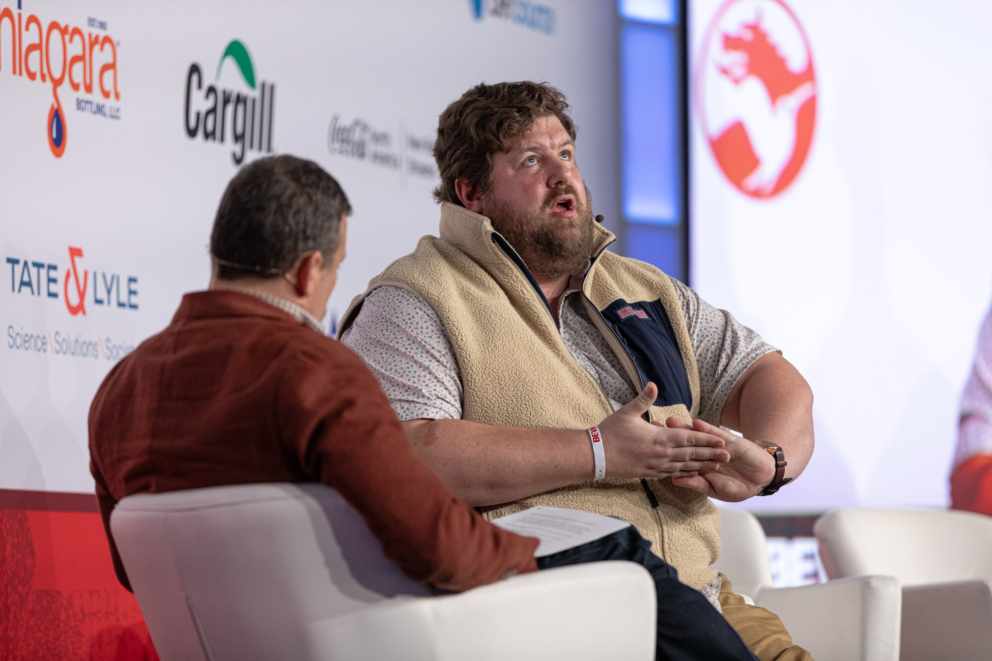 Man in a tan vest talks and gestures while seated across from a man in a red jacket.