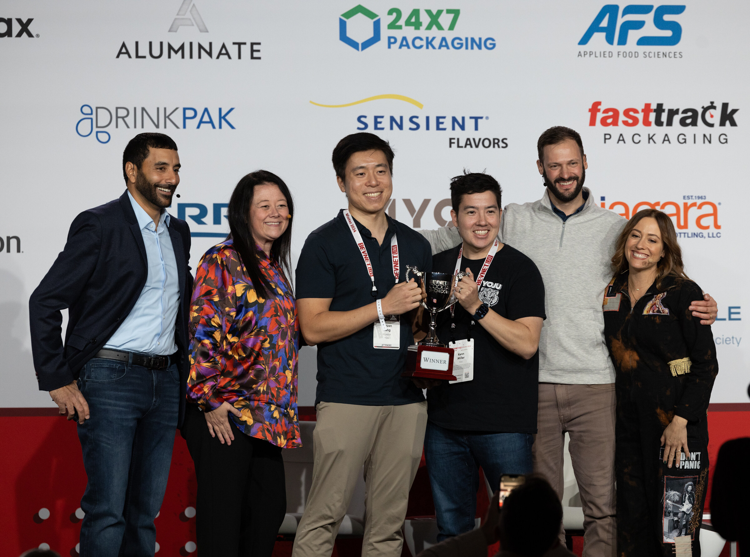 Six people stand together on stage, several holding a trophy, in front of a sponsor wall.