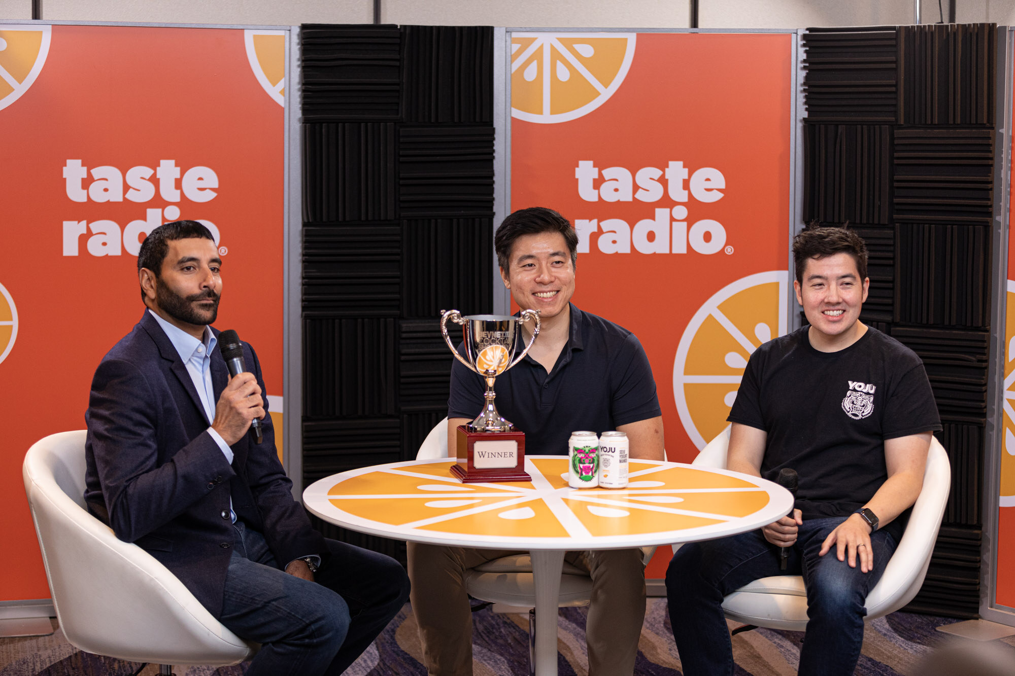 A host in a navy suit and two men in dark shirts sit around a round table with a winner trophy and two cans in front of a Taste Radio backdrop.