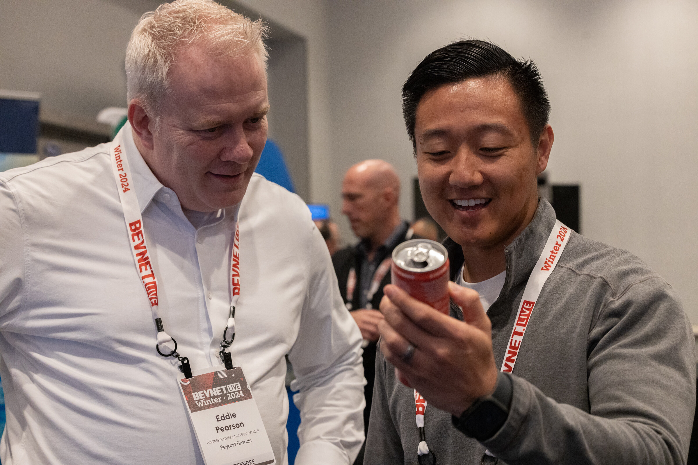 A man in a white shirt and a smiling man in a gray pullover look at a red can.