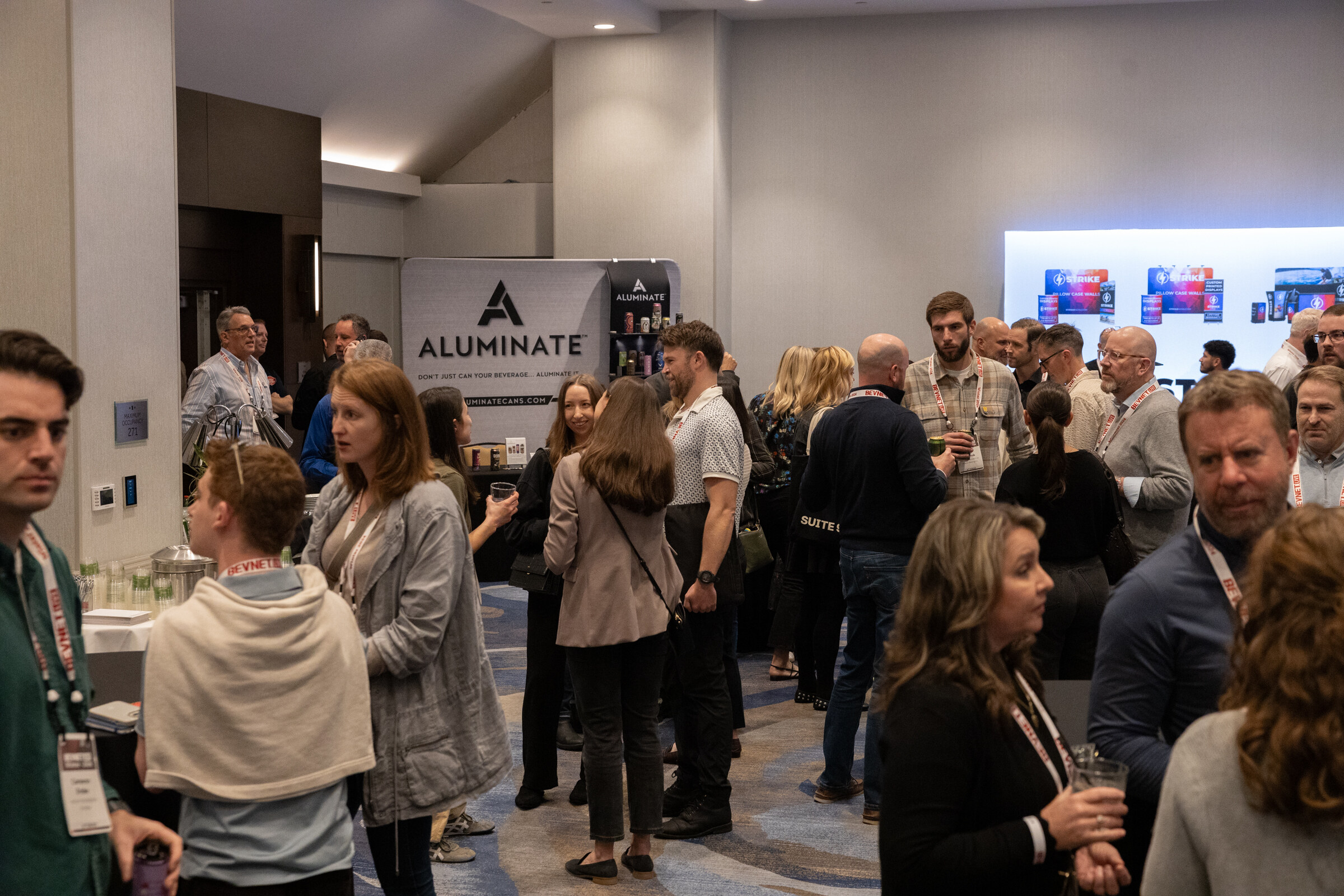 Crowd of attendees standing in a room with a banner for Aluminate and a display of cans in the background.