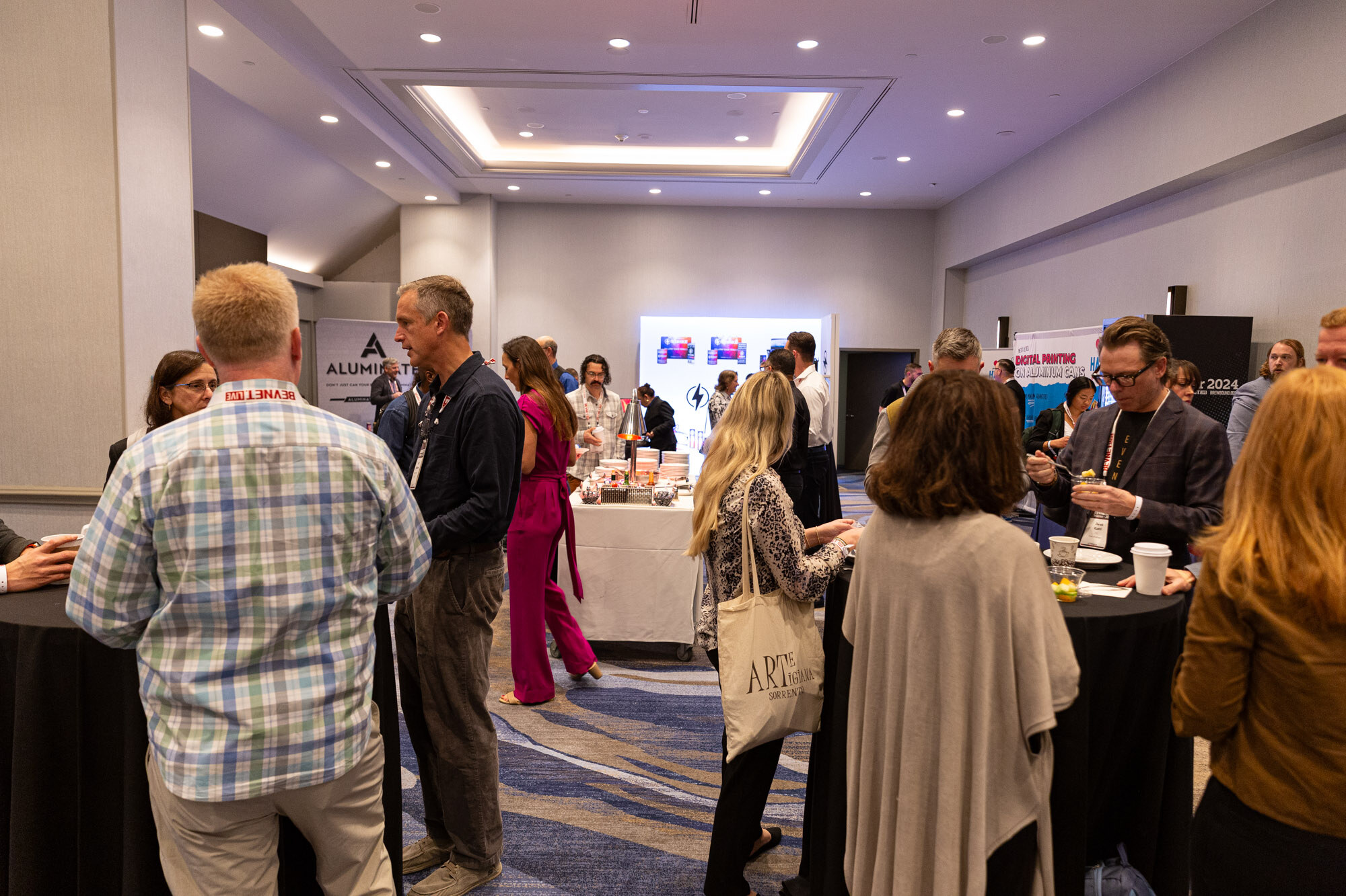People stand and talk around tall tables in a meeting room with bright displays at the far end.