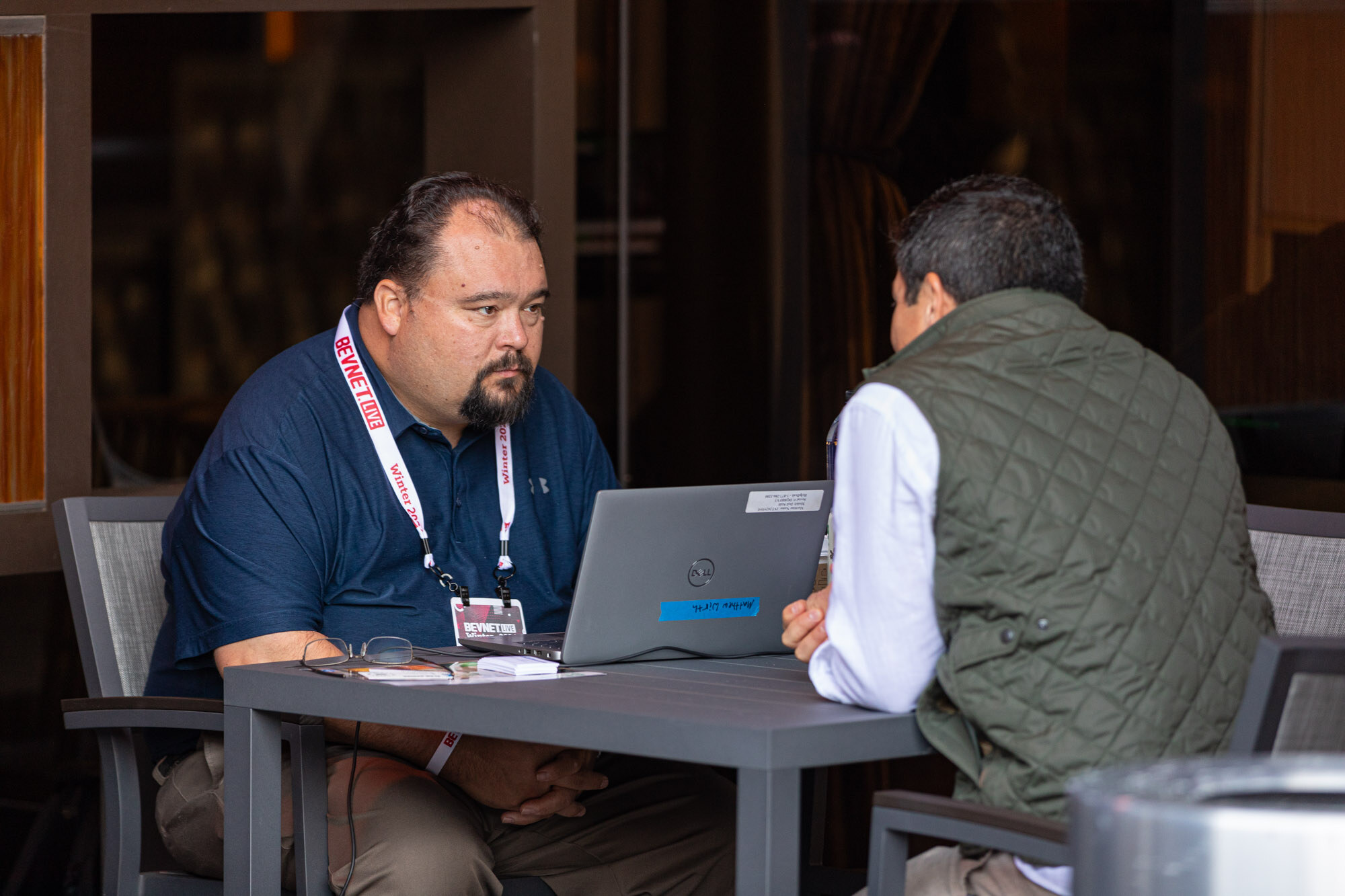 Two men sit across a table with a laptop and a lanyard badge on one man in a navy shirt.