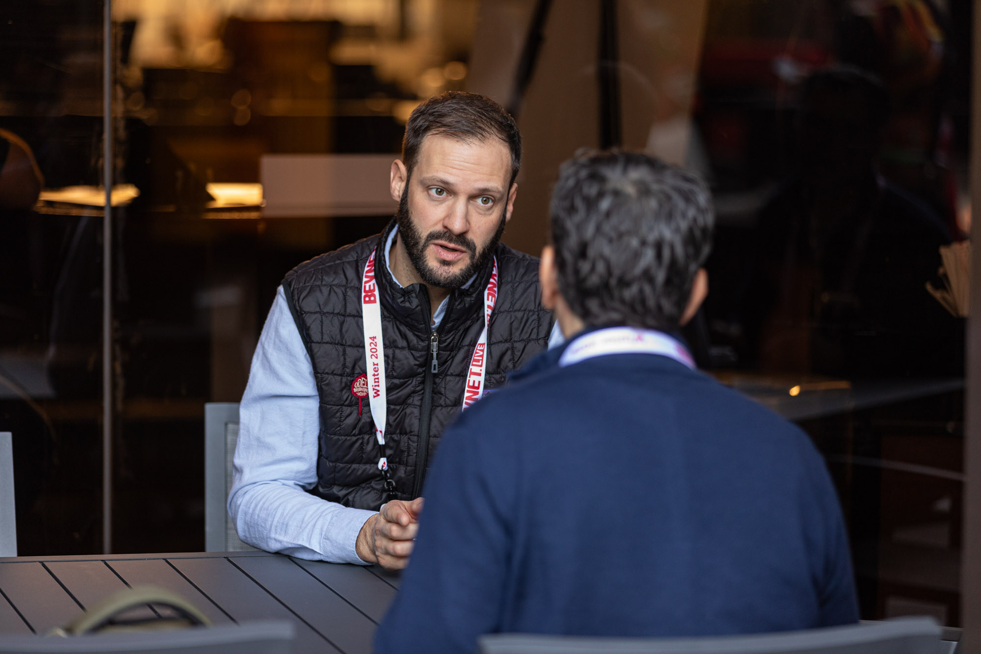 Bearded man in a black vest talks across the table while another man in navy sits with his back to the camera.