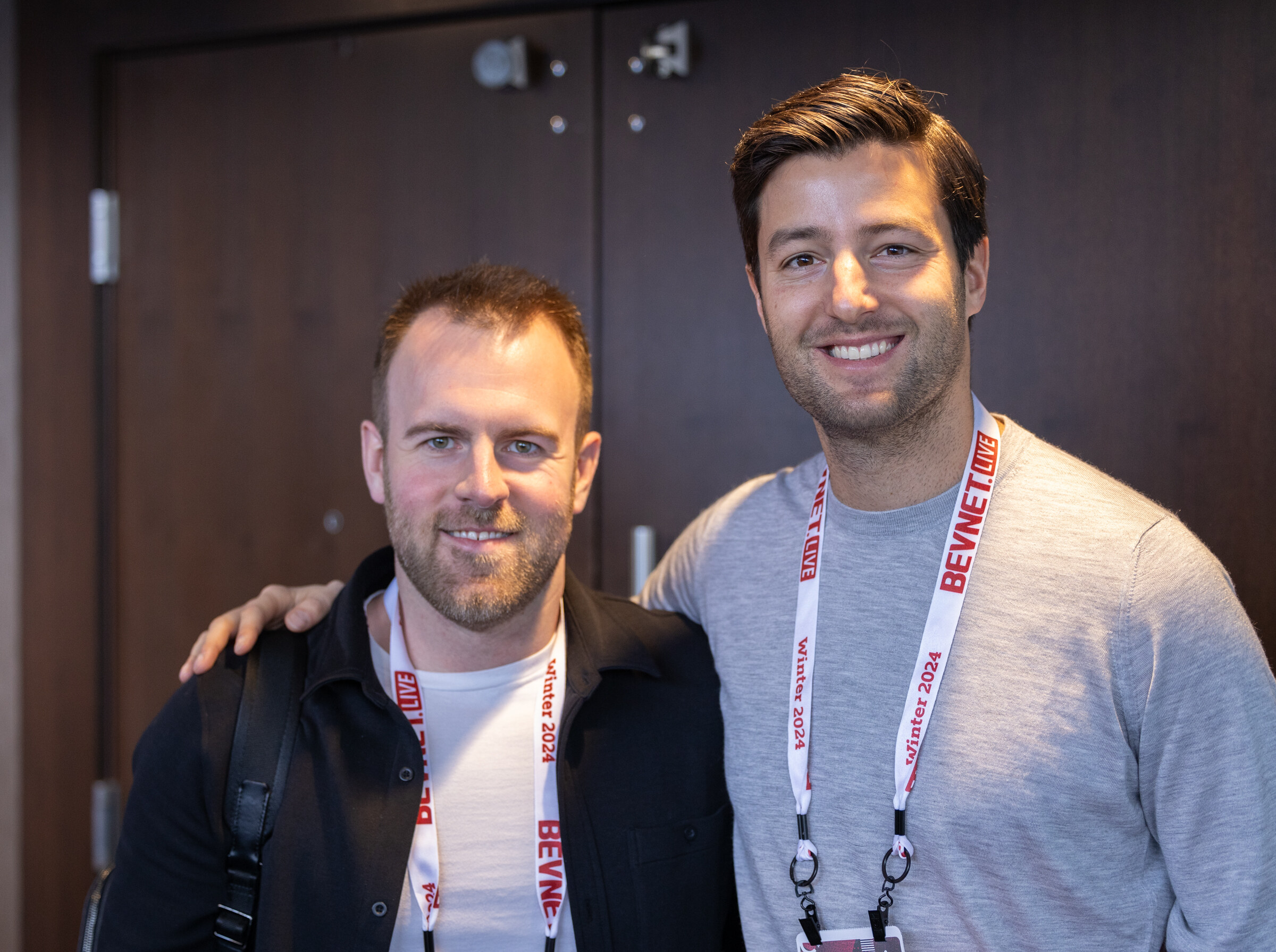 Two smiling men with BevNET Live Winter 2024 lanyards lean together in front of a wooden door.