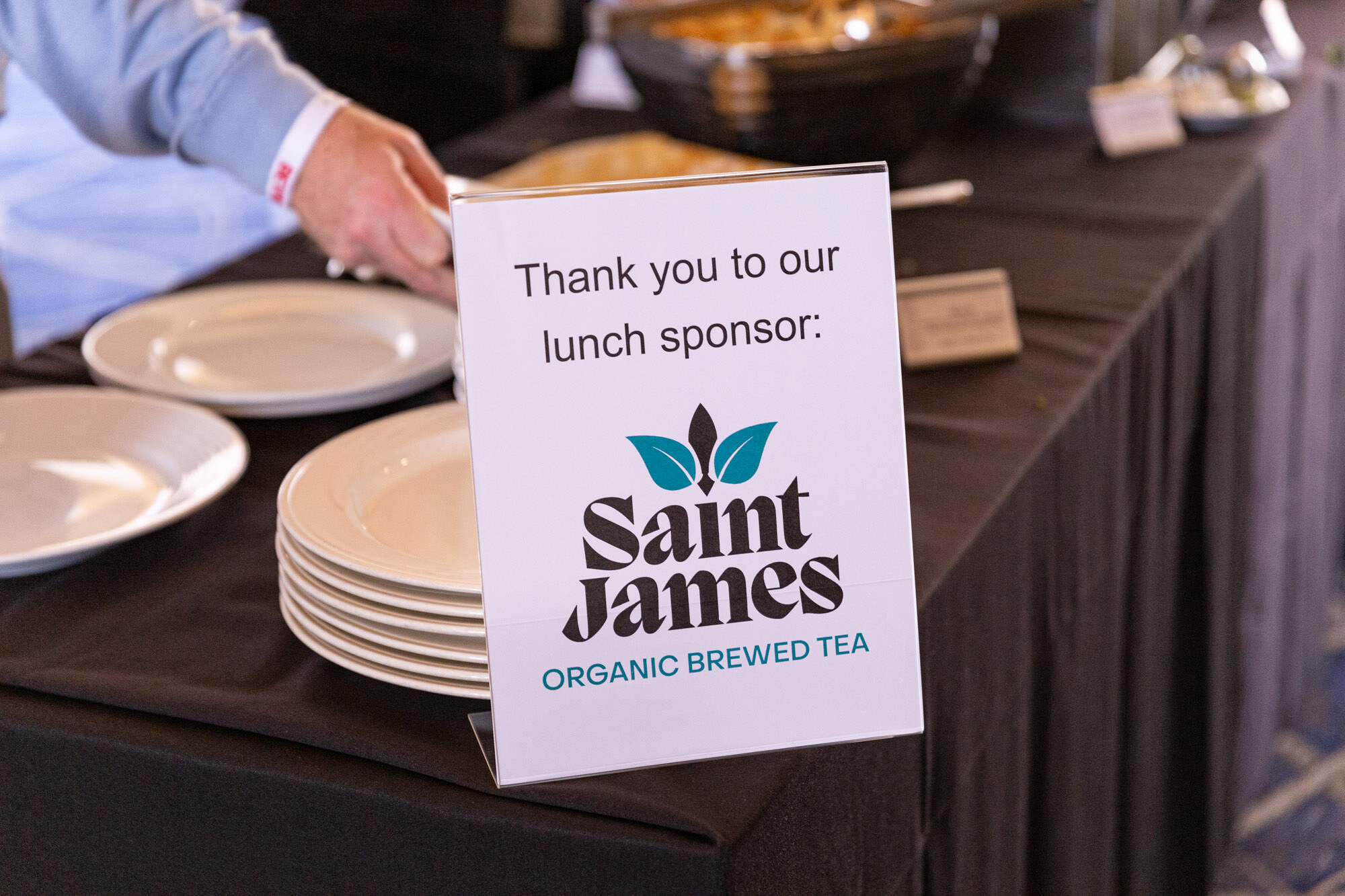 White sign reading Thank you to our lunch sponsor: Saint James Organic Brewed Tea next to a stack of plates.