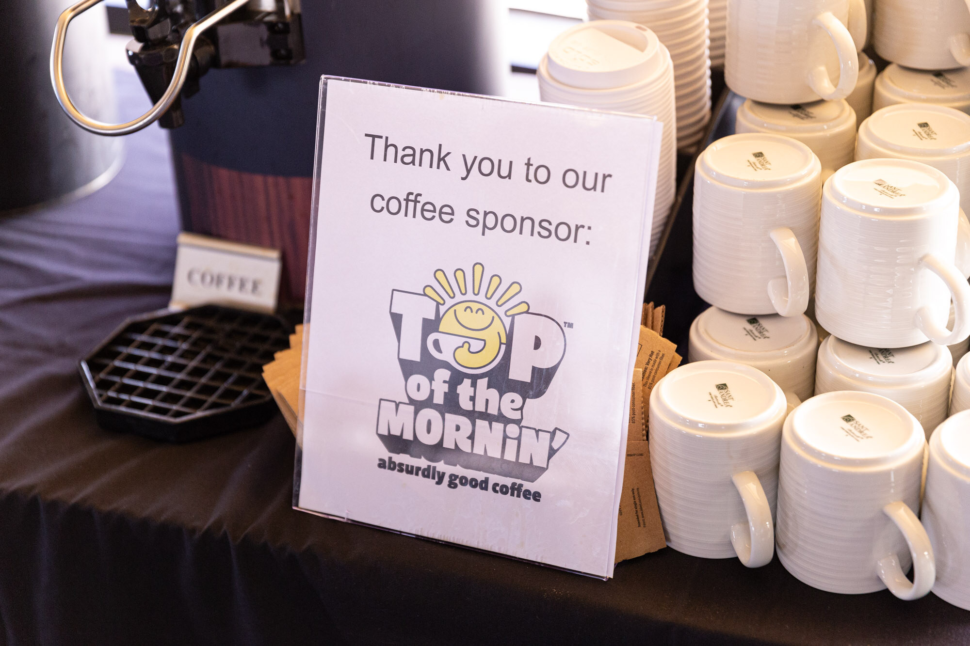 White sign reading Thank you to our coffee sponsor: Top of the Mornin' next to a stack of coffee cups.