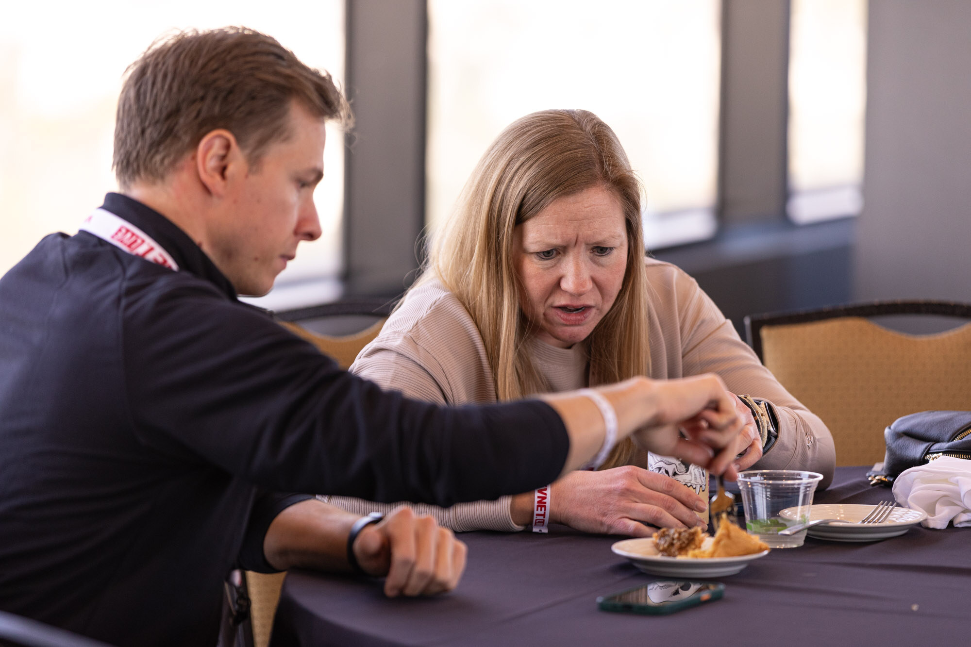 A man in a dark shirt and a woman in a beige sweater lean over a plate on a round table.