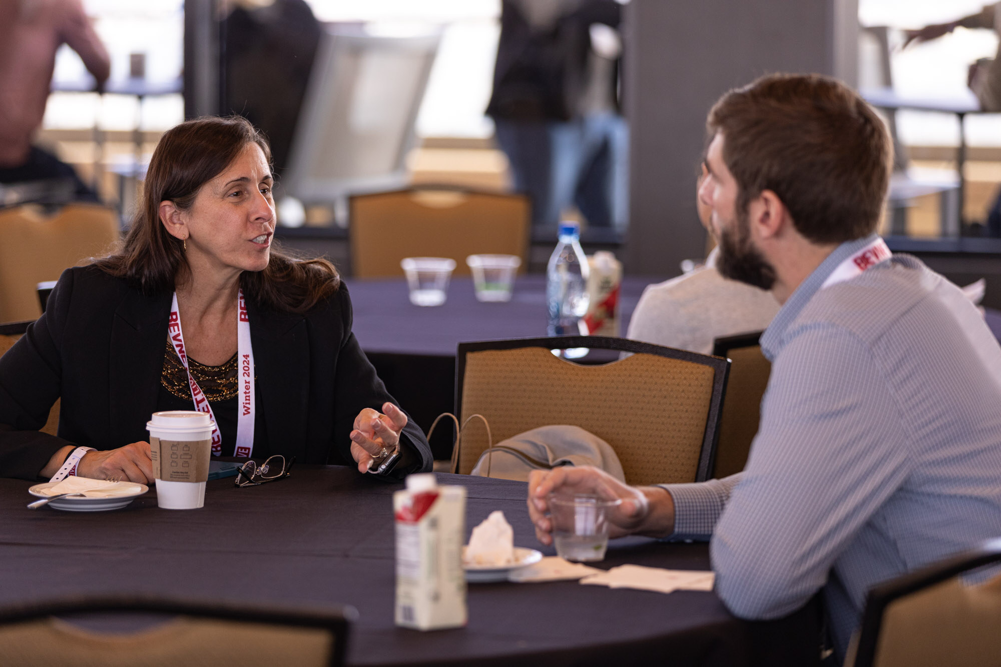 A woman with a lanyard talks with a bearded man at a table with a coffee cup and a carton.