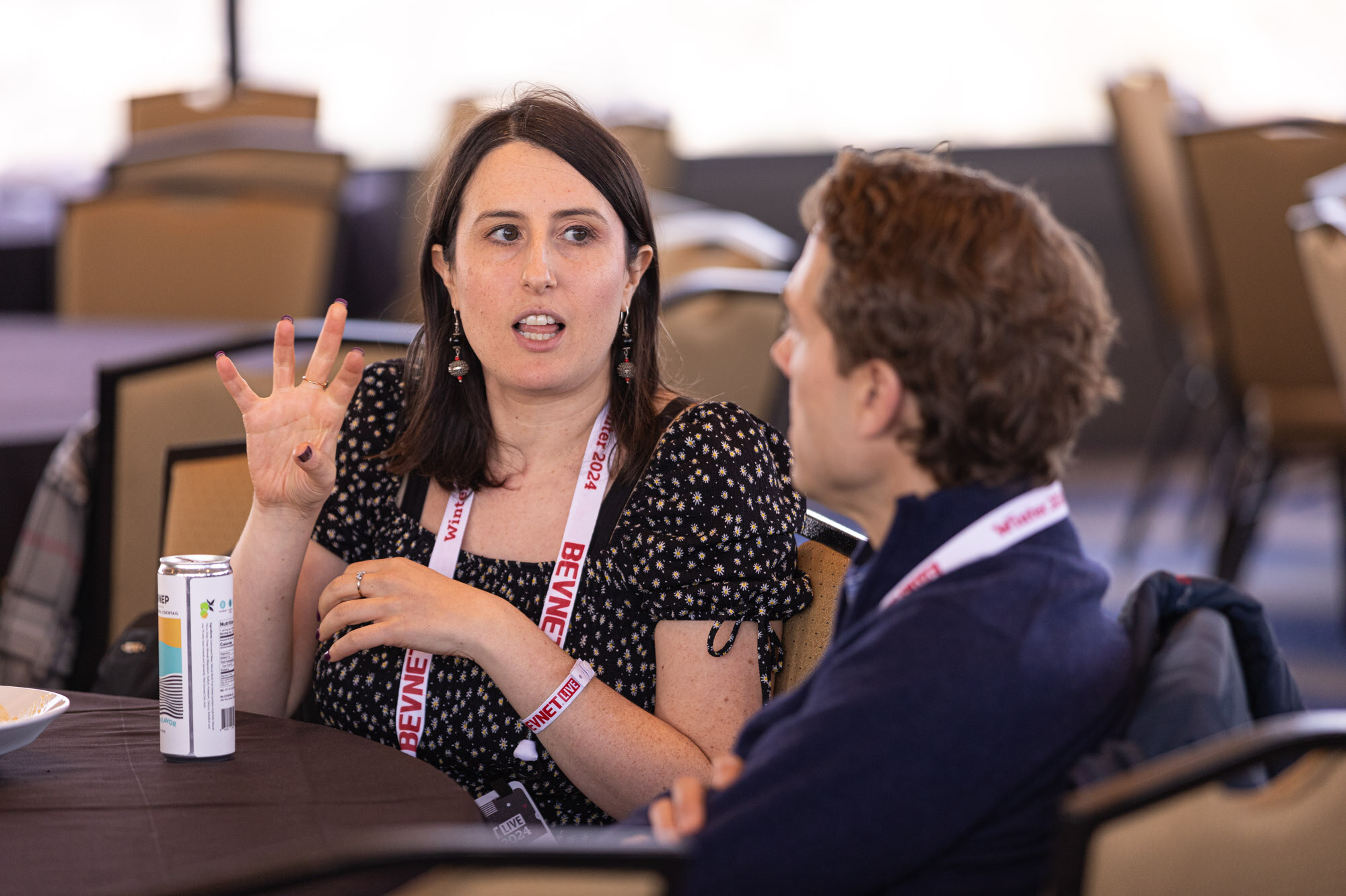 Woman in a black patterned dress holds up four fingers while talking to a man next to a can on the table.