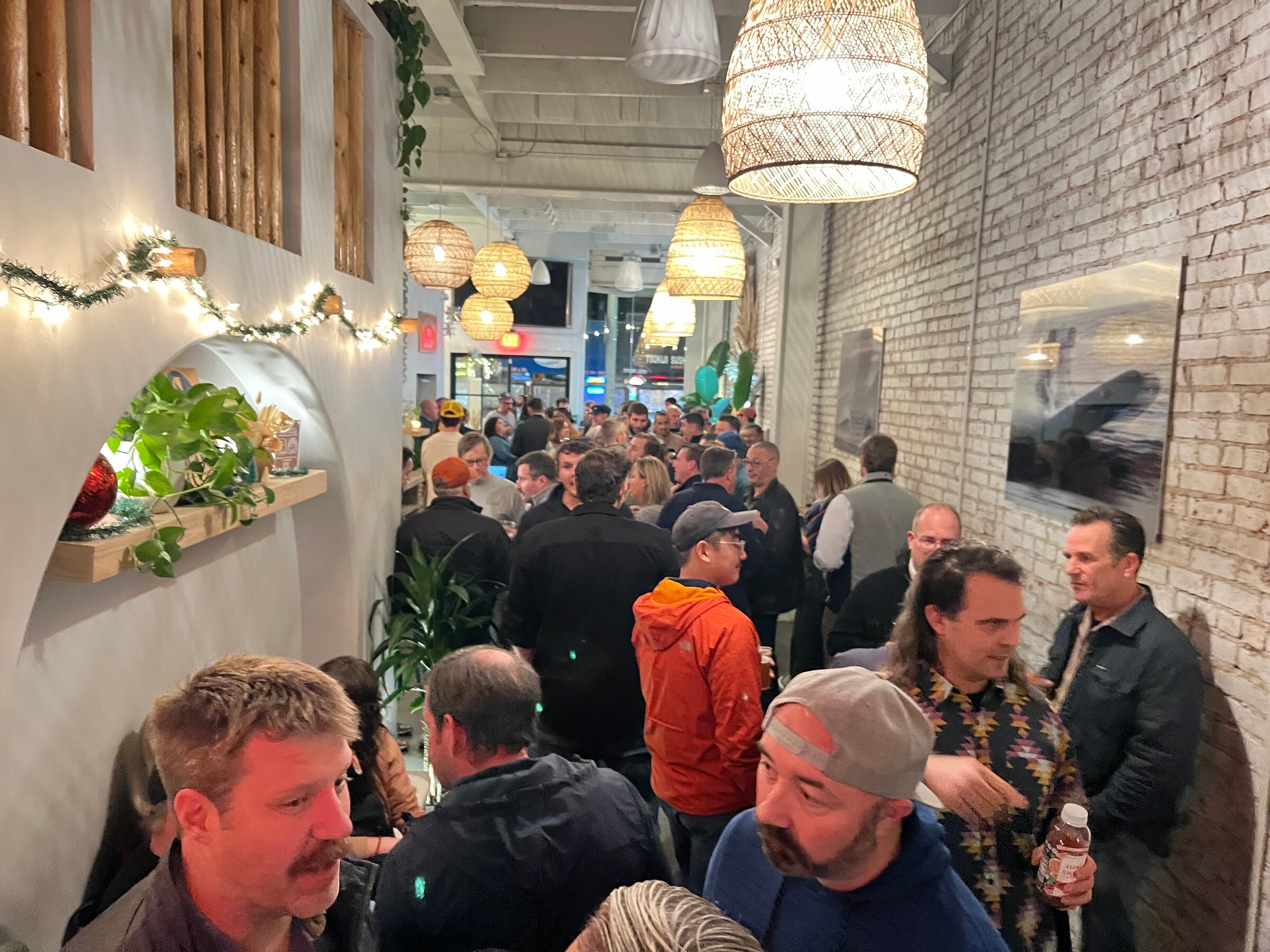 Packed narrow room with brick walls, string lights and a bar in the background; people stand close together in the foreground.