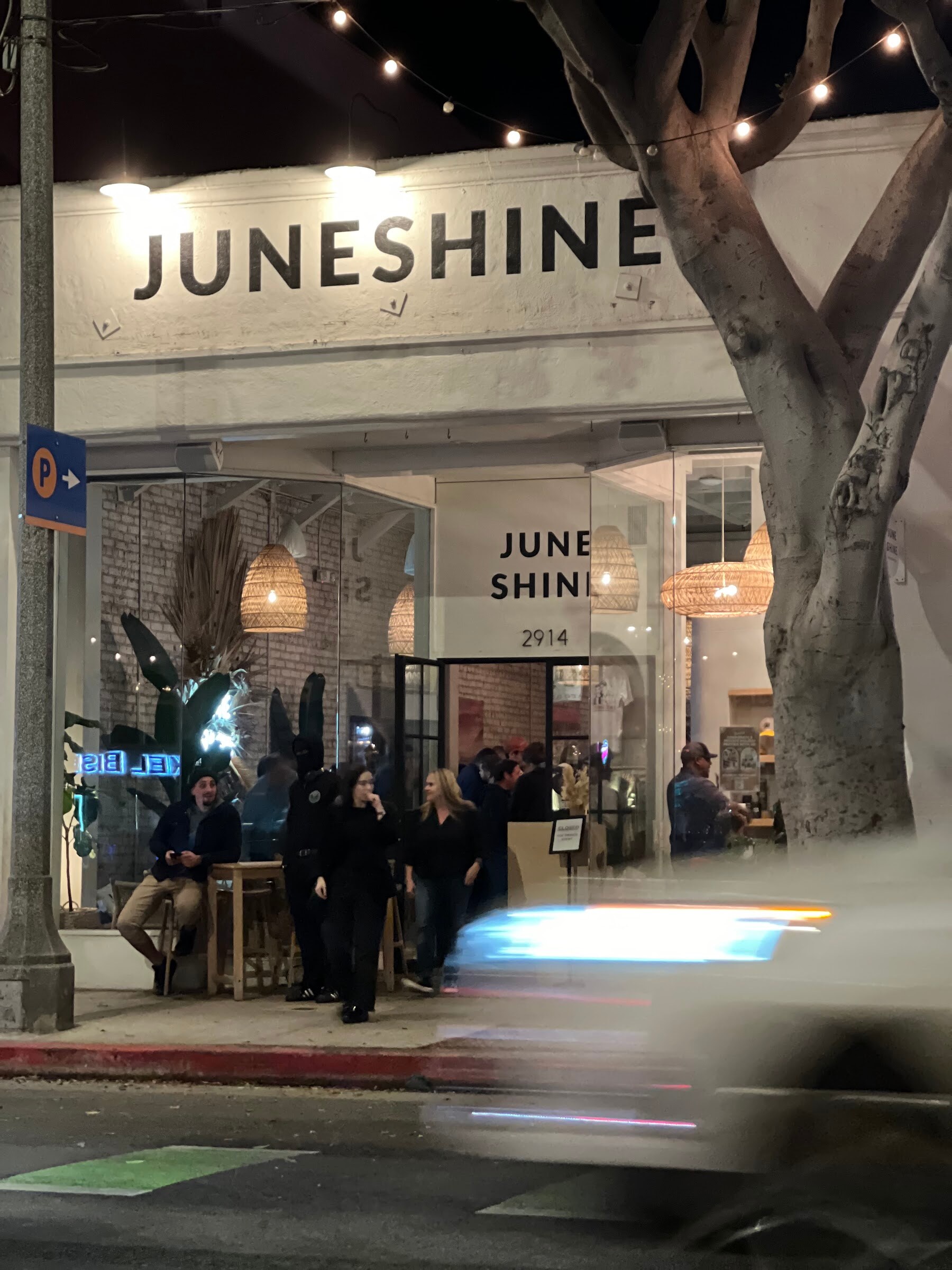 Nighttime view of a storefront with the word JUNESHINE above the entrance and people gathered inside.