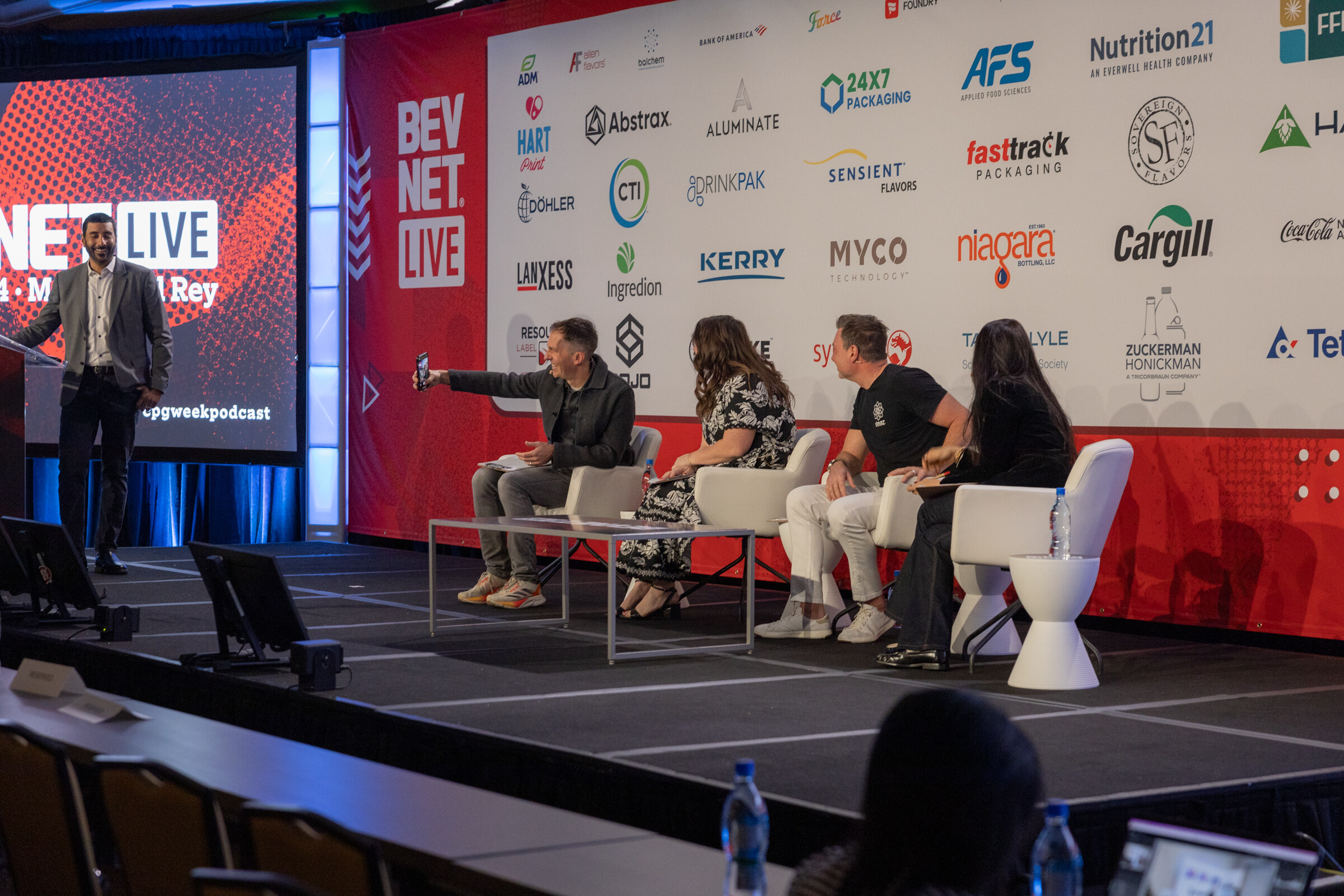 Wide shot of a man standing at left and four judges seated in white chairs on stage in front of a BevNET Live backdrop.