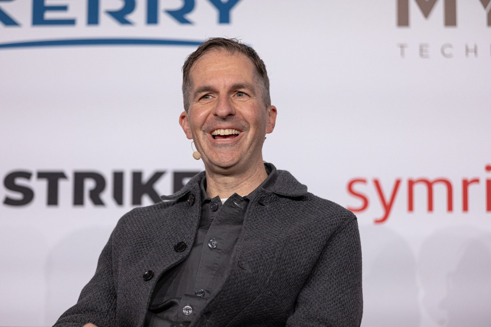 Smiling man in a dark gray jacket sits in front of a sponsor wall.