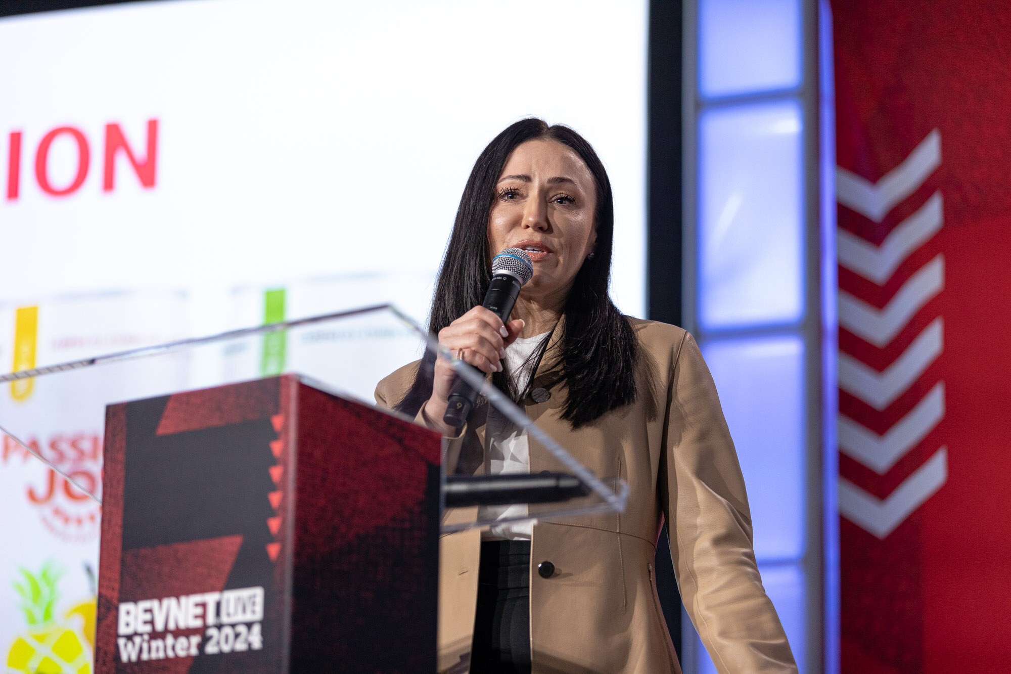 Woman with long dark hair in a beige jacket speaks at a podium in front of a slide.