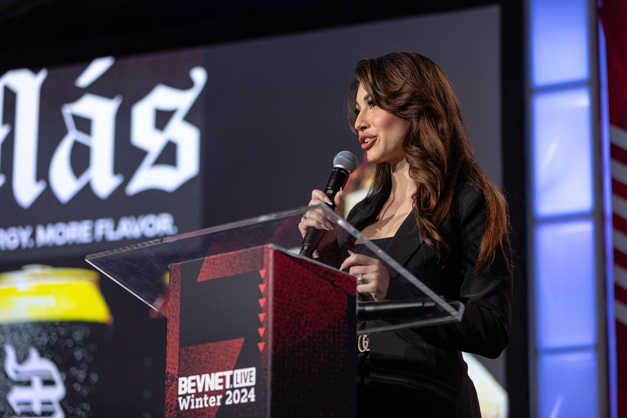 Woman with wavy brown hair in a black jacket speaks at a podium that reads BevNET Live Winter 2024.