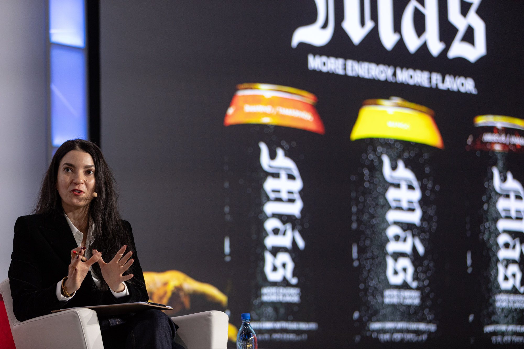 Woman in a black blazer gestures while seated in front of a screen showing dark cans labeled Mas.