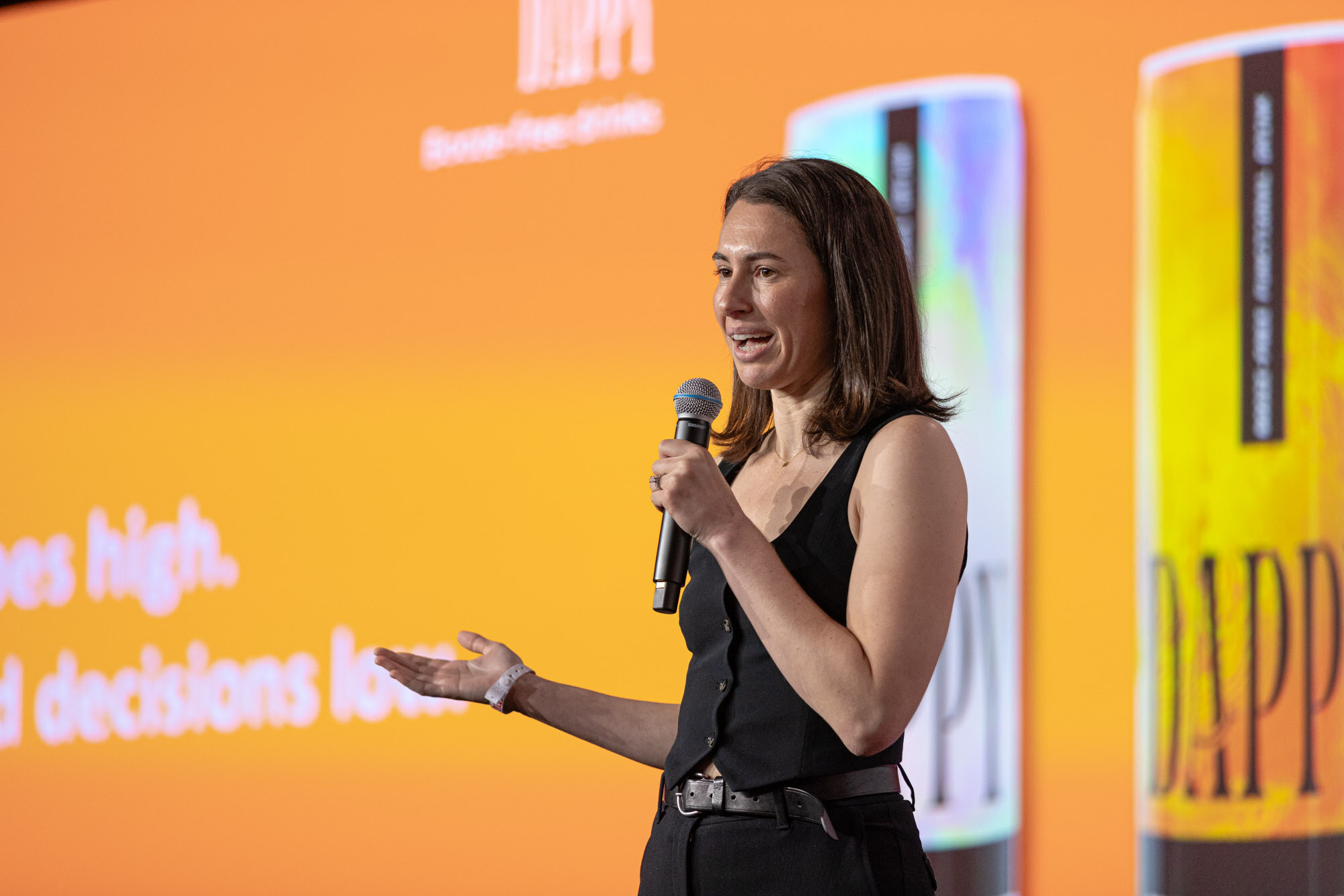 Woman in a black vest holds a microphone and gestures in front of an orange screen showing tall Dappy cans.