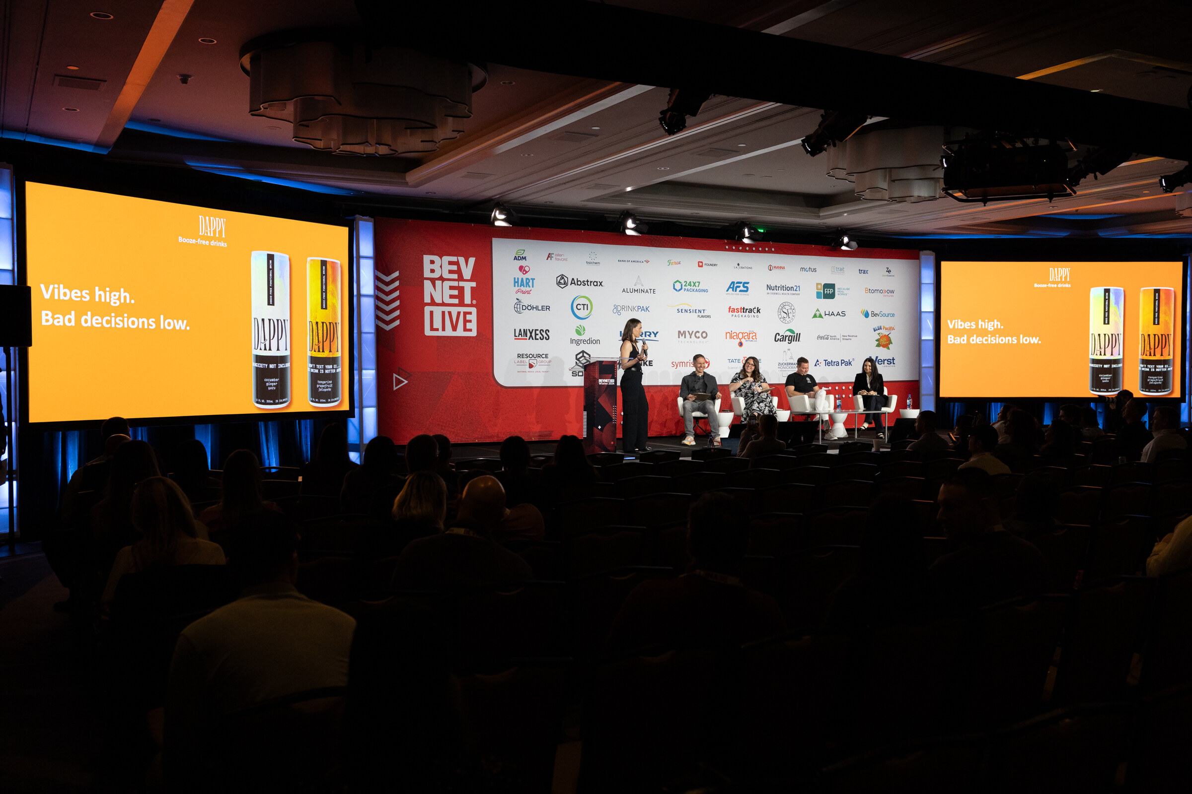 Dark ballroom with a lit stage, a presenter at a podium and orange screens that read Vibes high bad decisions low.