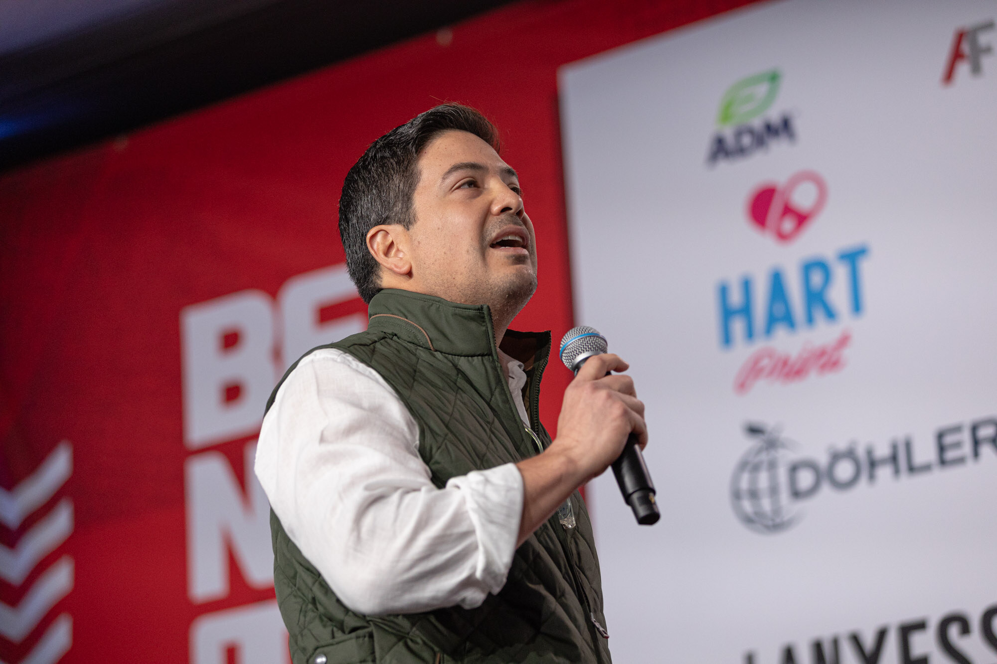 Man in an olive vest over a white shirt holds a microphone and looks up in front of a red BevNET Live backdrop with sponsor logos.