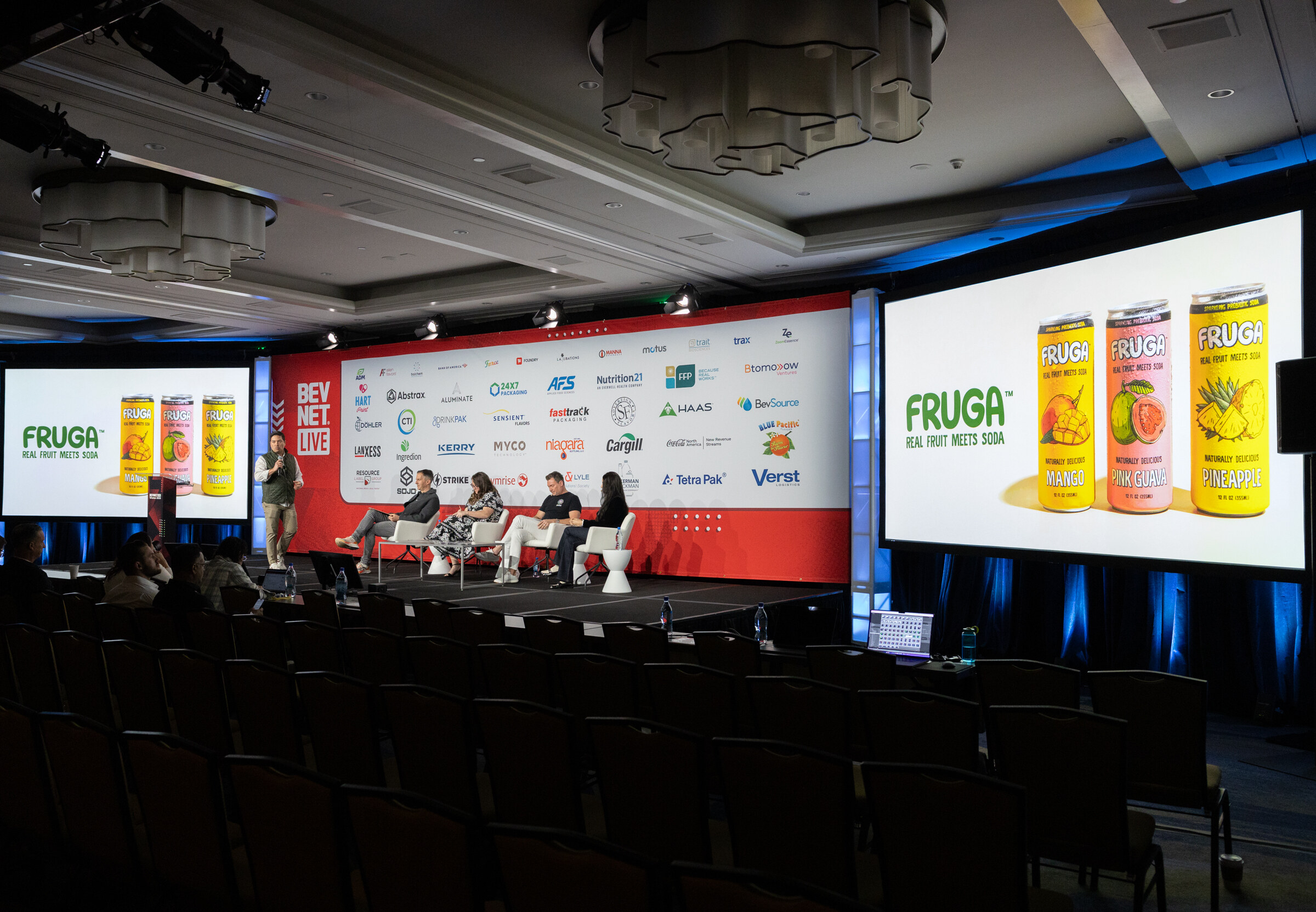 Dark ballroom with a lit stage and large screens showing Fruga real fruit melts soda cans.