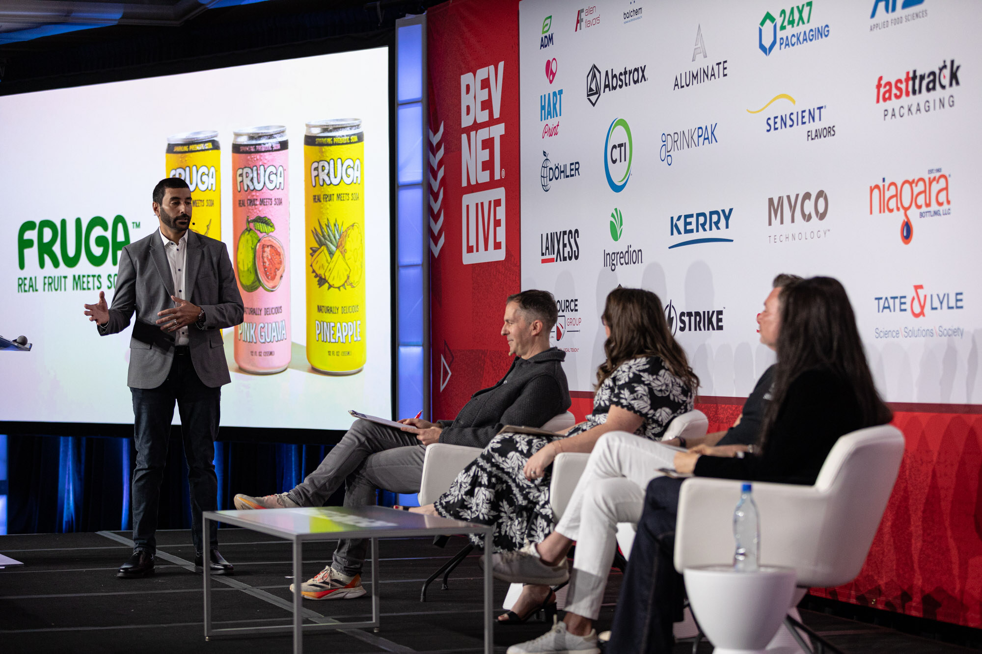 Man in a gray blazer stands left of the judges in front of a screen with Fruga real fruit melts soda cans.