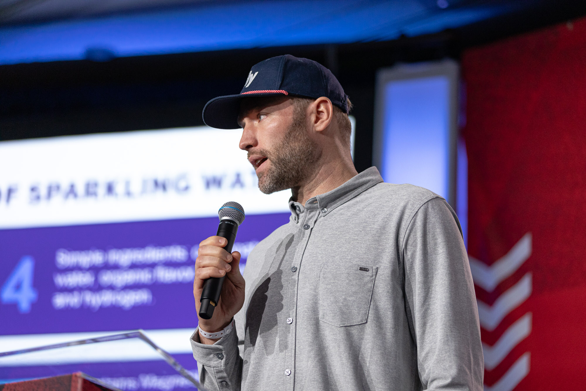 Man in a navy cap and gray shirt holds a microphone in front of a purple slide that mentions sparkling water.