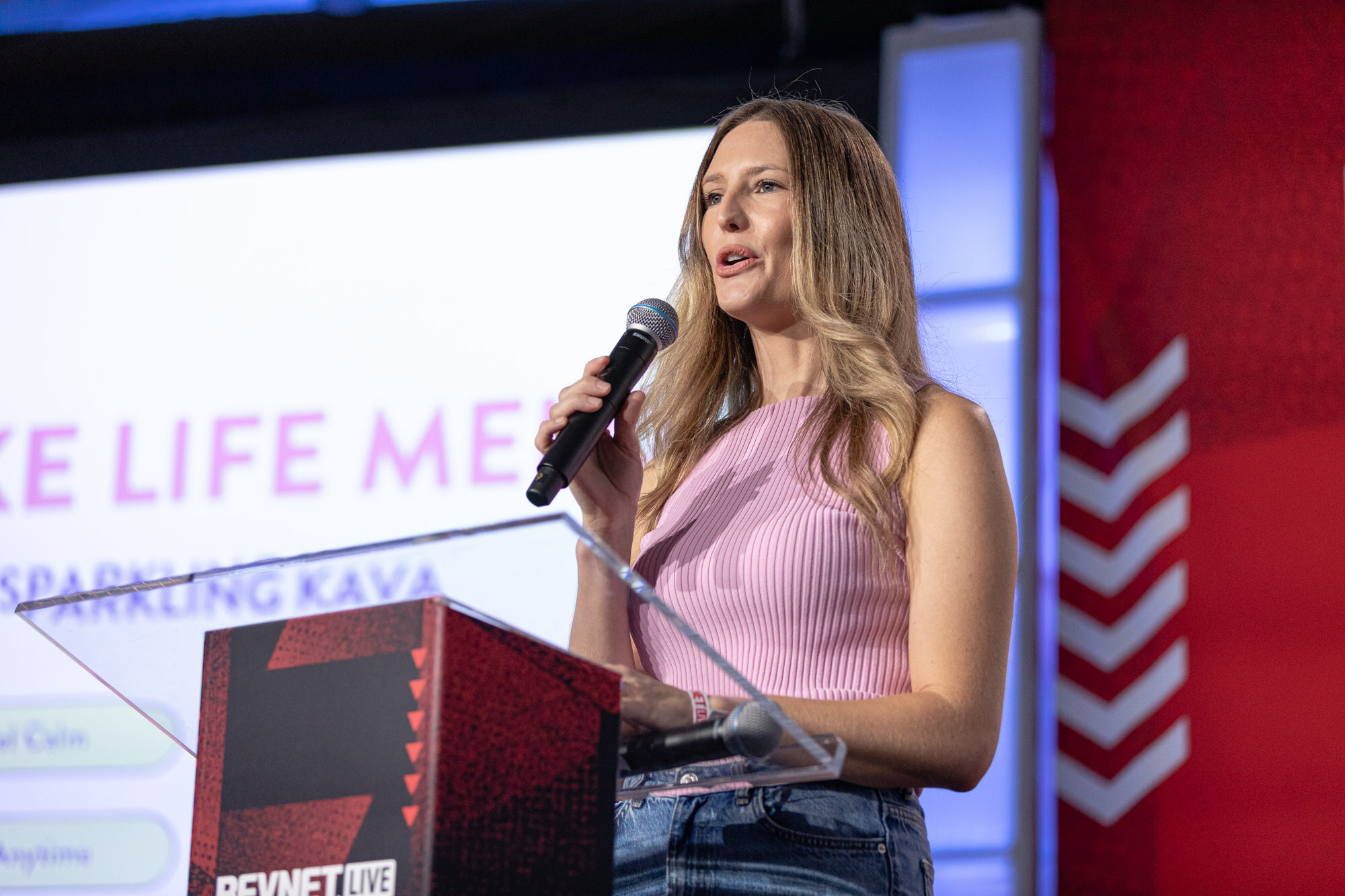 Woman in a pink top and jeans speaks into a microphone at a podium in front of a slide reading Make Life Melo.