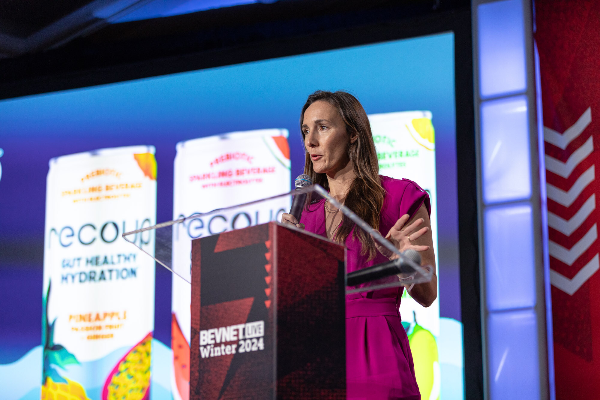 Woman in a magenta dress speaks at a podium in front of a large screen showing recoup gut healthy hydration cans.
