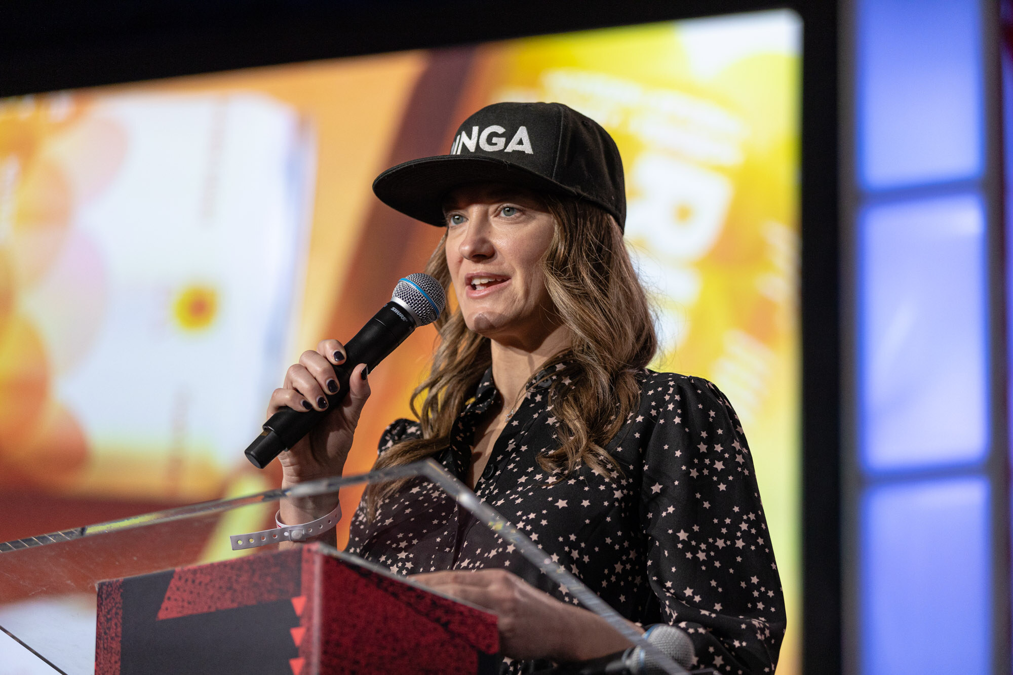 Woman in a cap and a polka-dot top speaks into a microphone behind a podium in front of an orange screen.