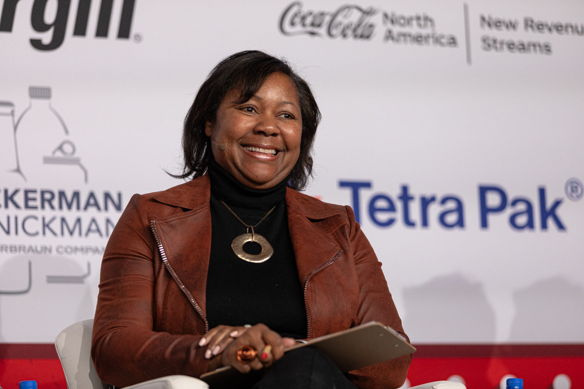 Smiling woman in a brown leather jacket holds a tablet while seated in front of a sponsor wall.
