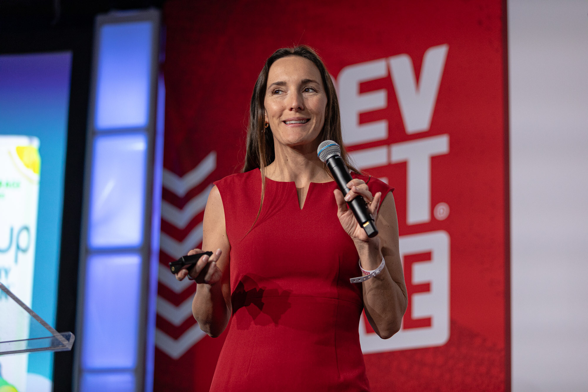Woman in a red dress holds a microphone and smiles on stage in front of a red BevNET Live backdrop.