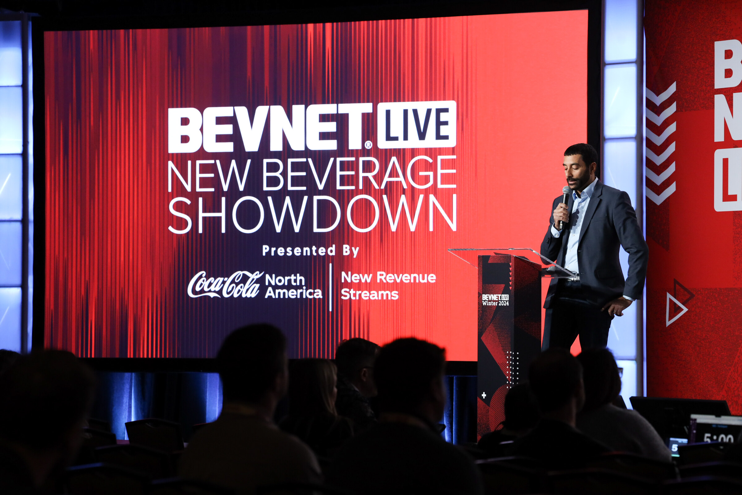 Man in a dark suit stands beside a podium next to a screen reading BevNET Live New Beverage Showdown presented by Coca-Cola North America.