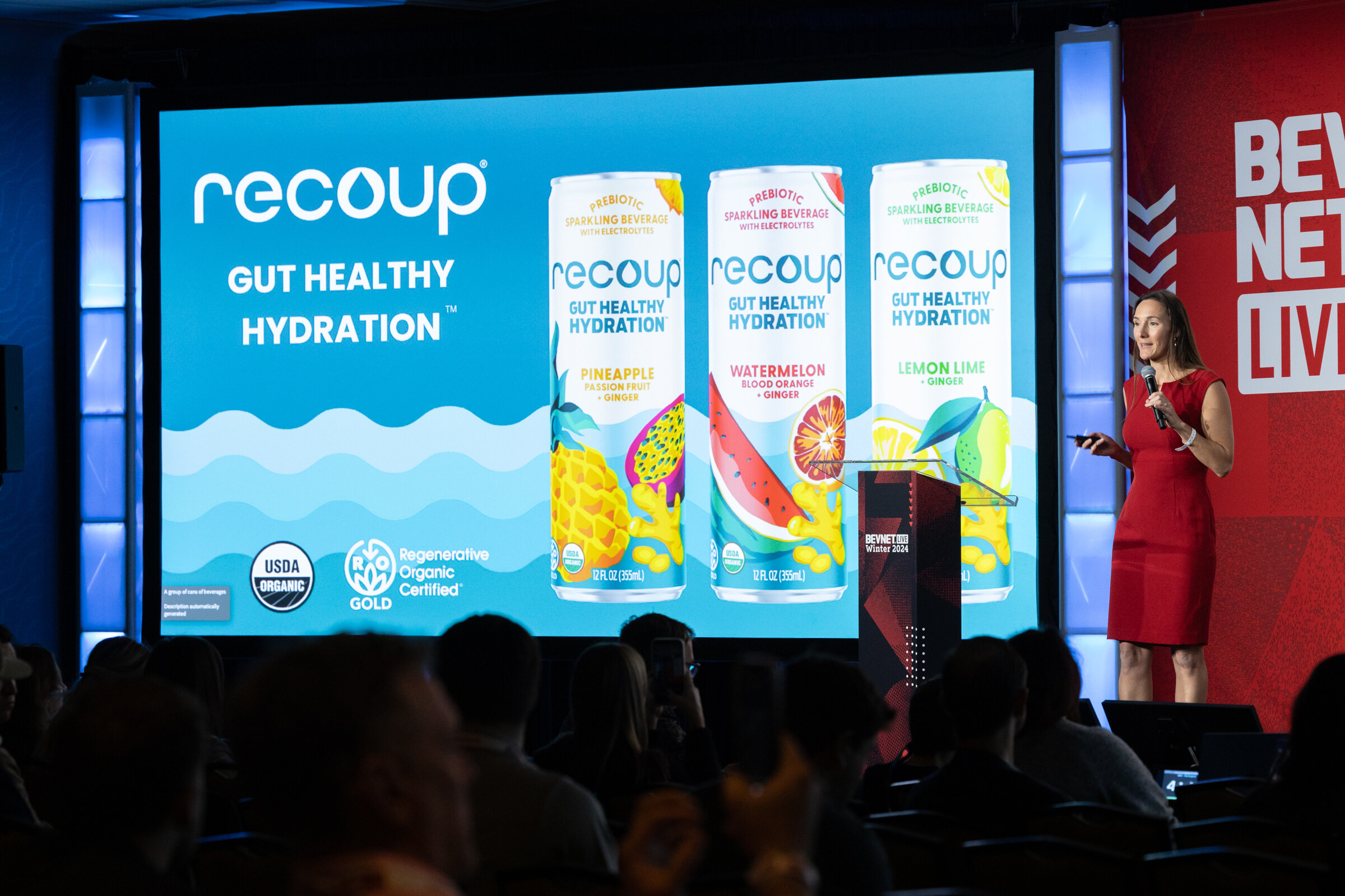 Woman in a red dress stands next to a podium in front of a screen showing three recoup cans.