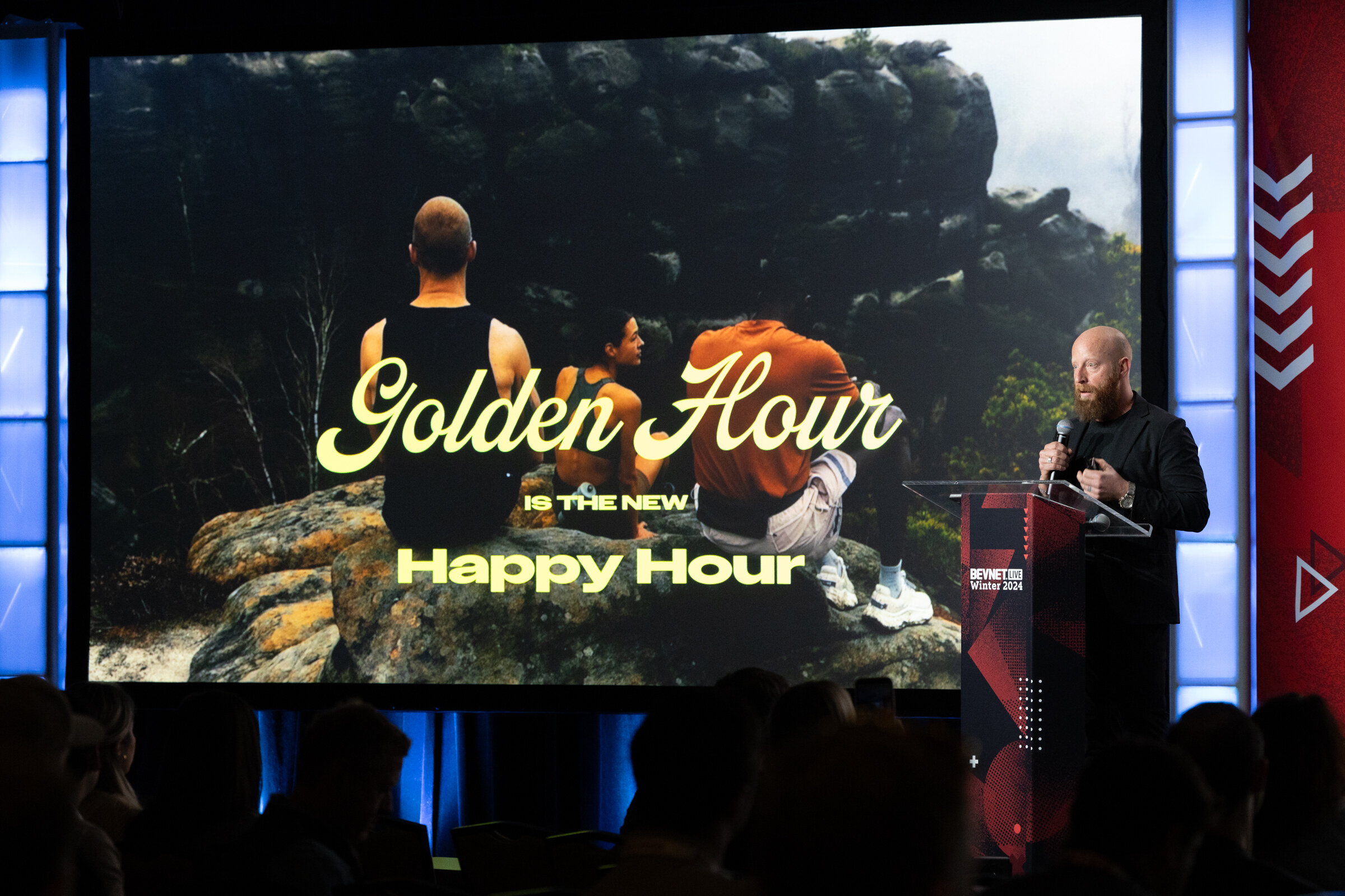 Man at a podium on the right of a large screen reading Golden Hour is the new Happy Hour.