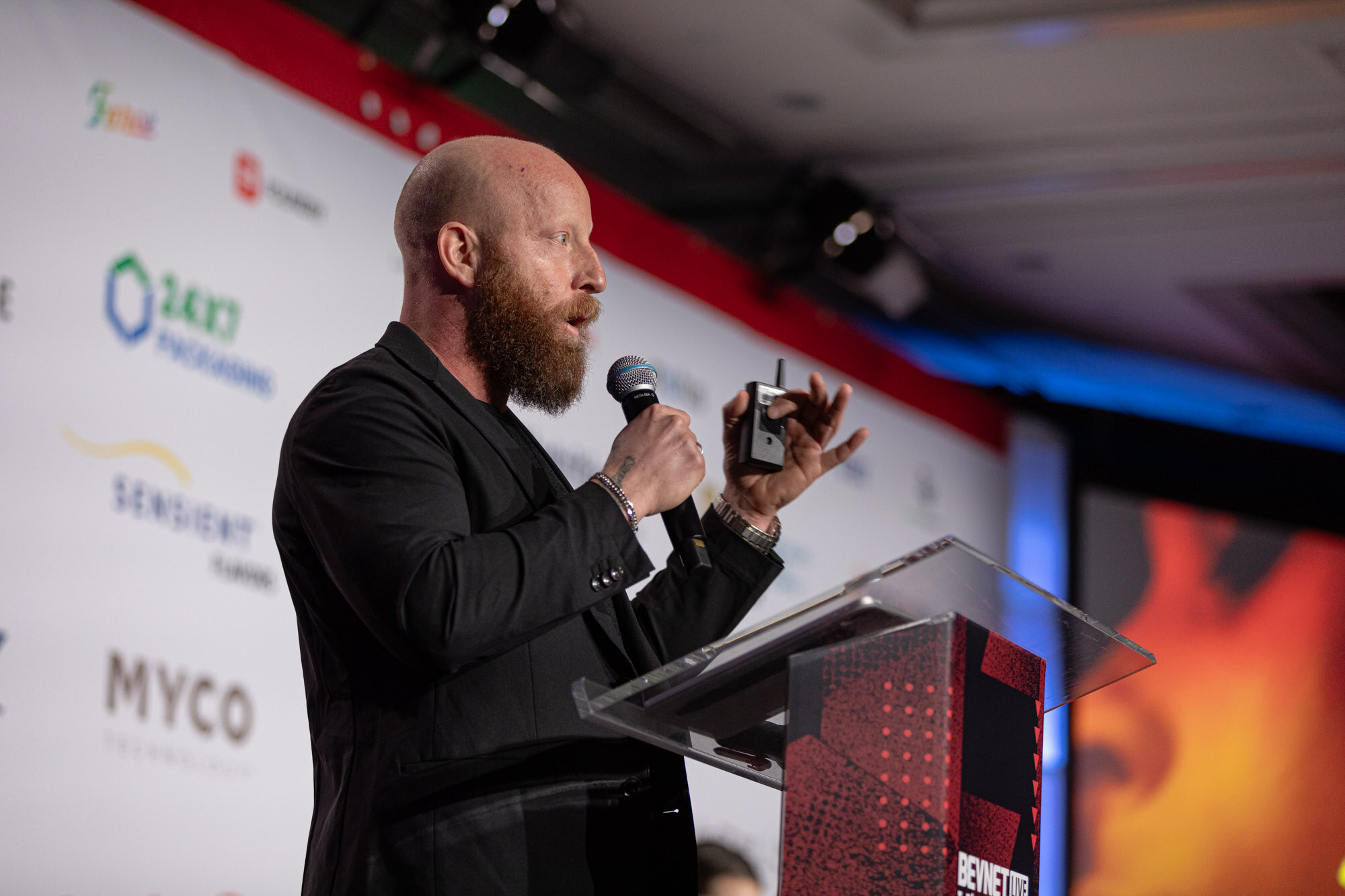 Bearded man in a black jacket speaks at a clear podium in front of a sponsor backdrop.