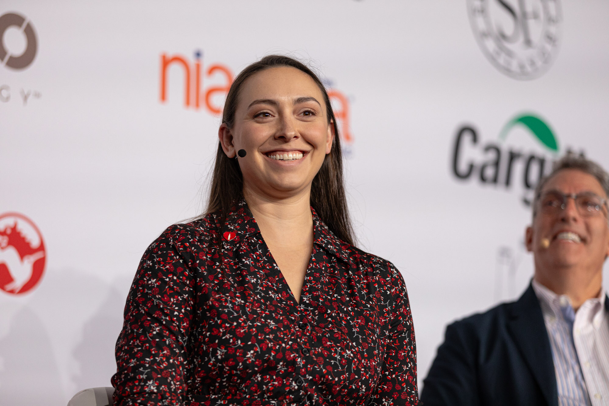 Smiling woman in a patterned blouse sits on stage next to a man leaning in laughing.