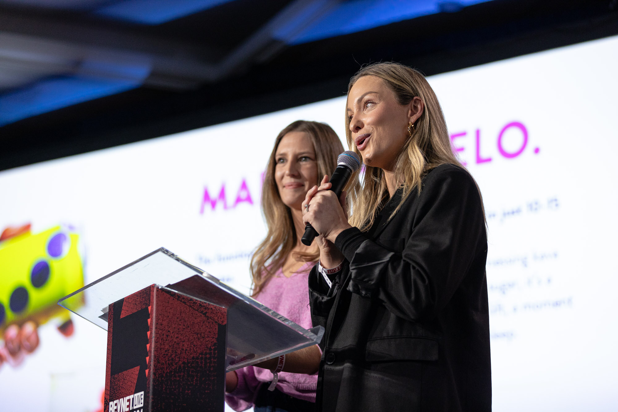 Two women stand together at a lectern in front of a screen reading MELO, one speaking into a microphone.