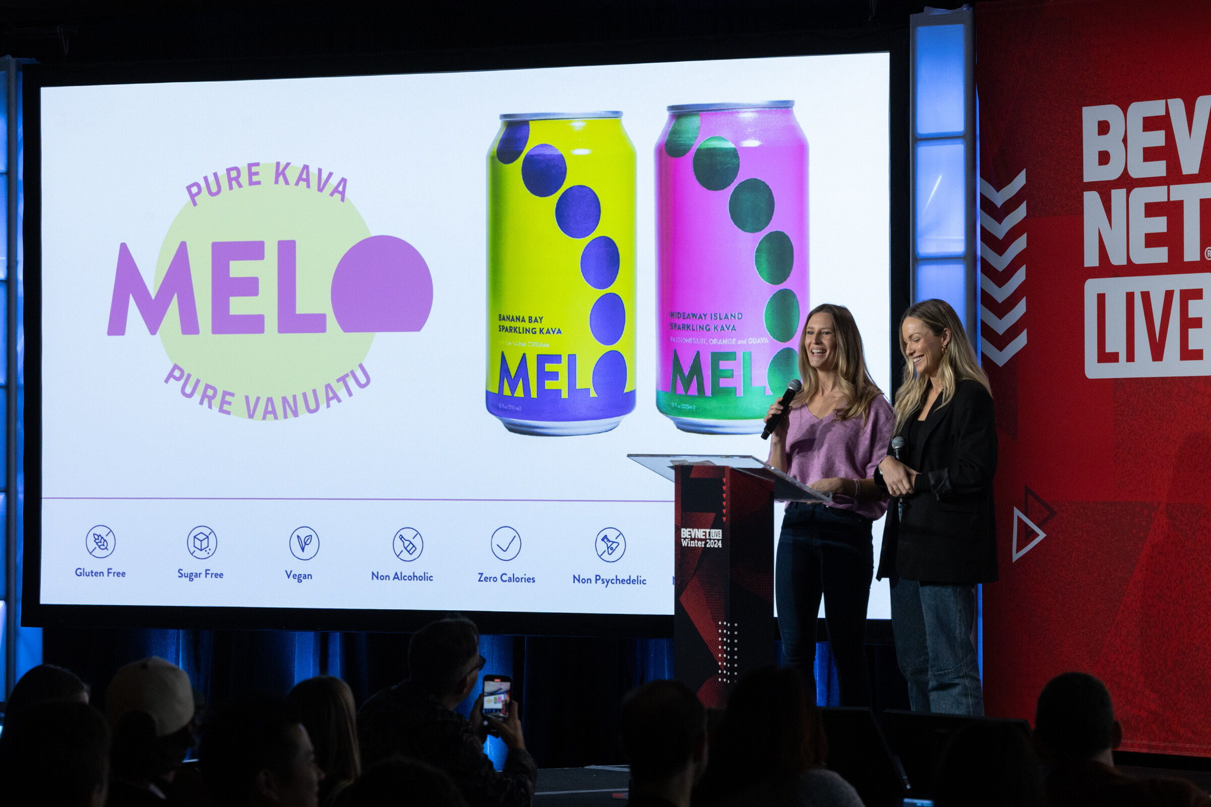 Two women stand at a podium in front of a screen with MELO pure kava cans in yellow and pink.