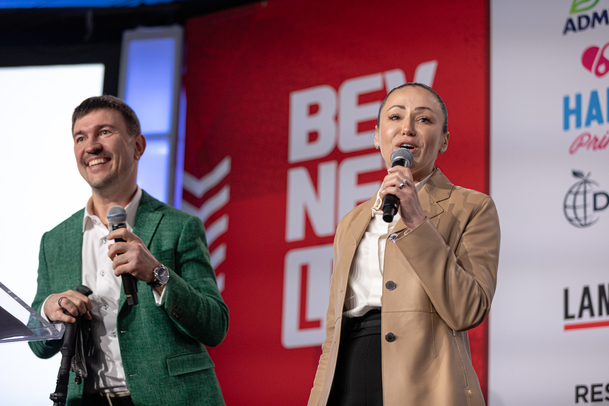 A man in a green blazer and a woman in a tan blazer hold microphones and smile on stage.