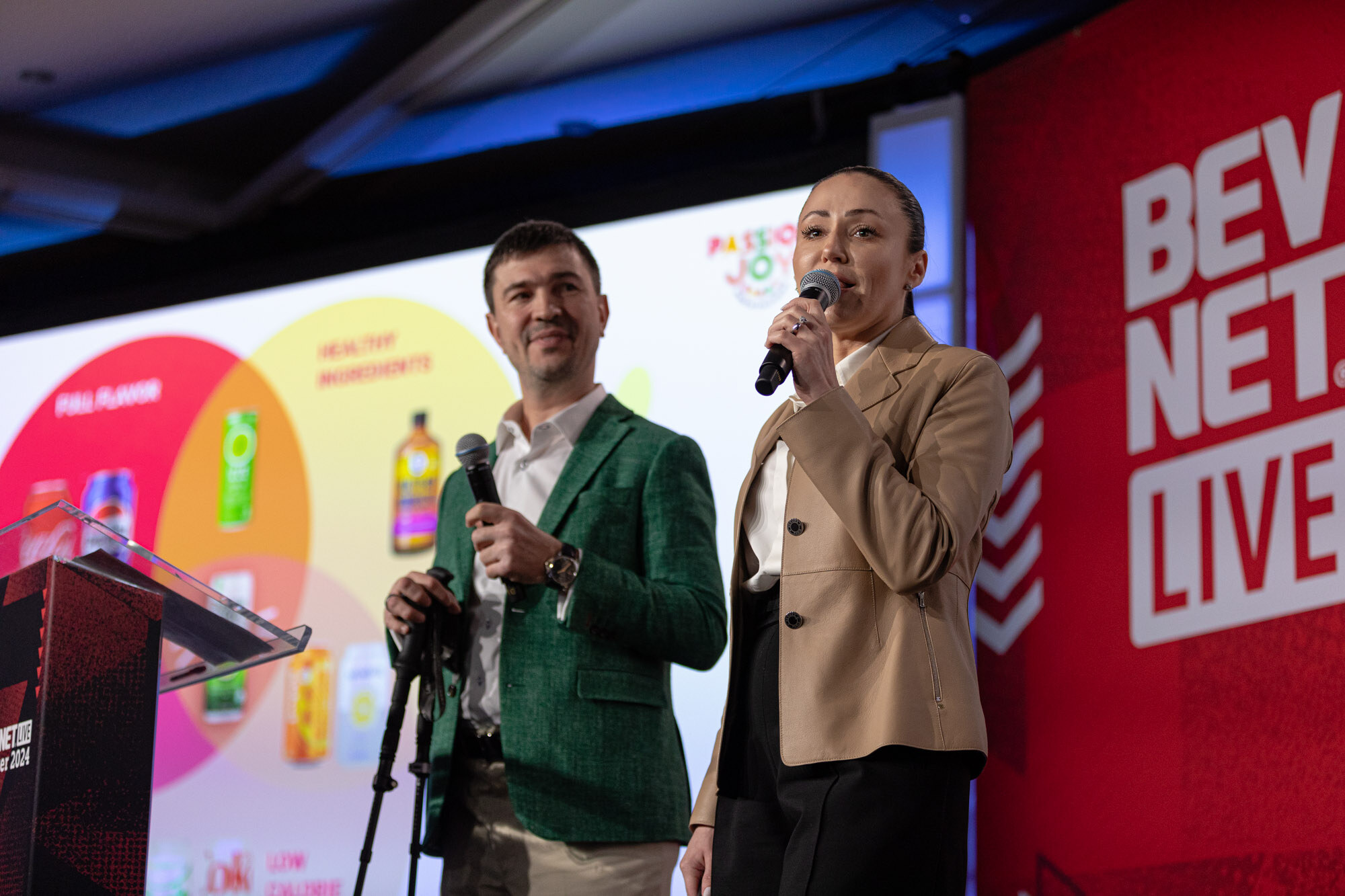 A man in a green blazer and a woman in a tan blazer stand at a podium in front of a colorful circle graphic.