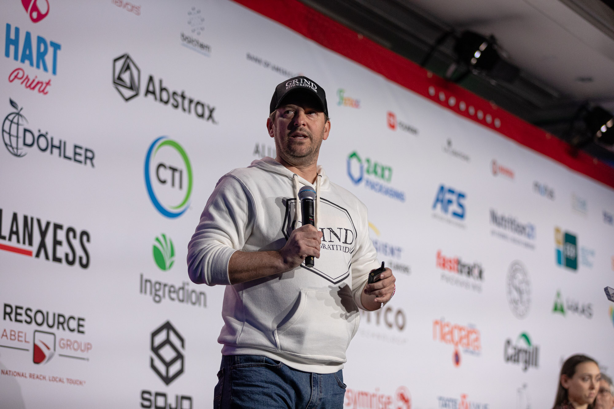 Man in a white hoodie and black cap holds a microphone at the stage in front of a sponsor wall.