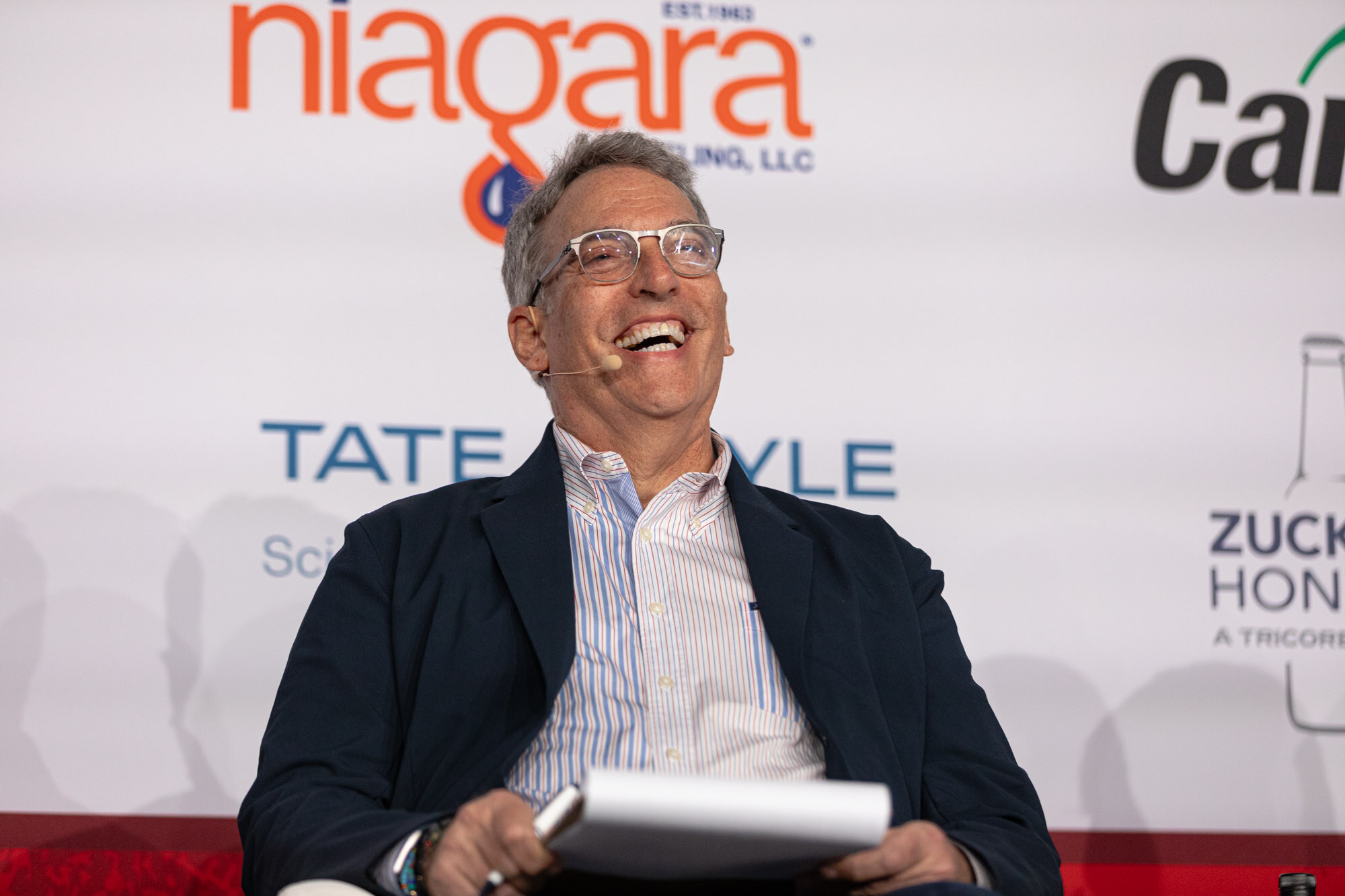 Man with glasses in a navy blazer laughs while holding a notepad in front of a sponsor wall.