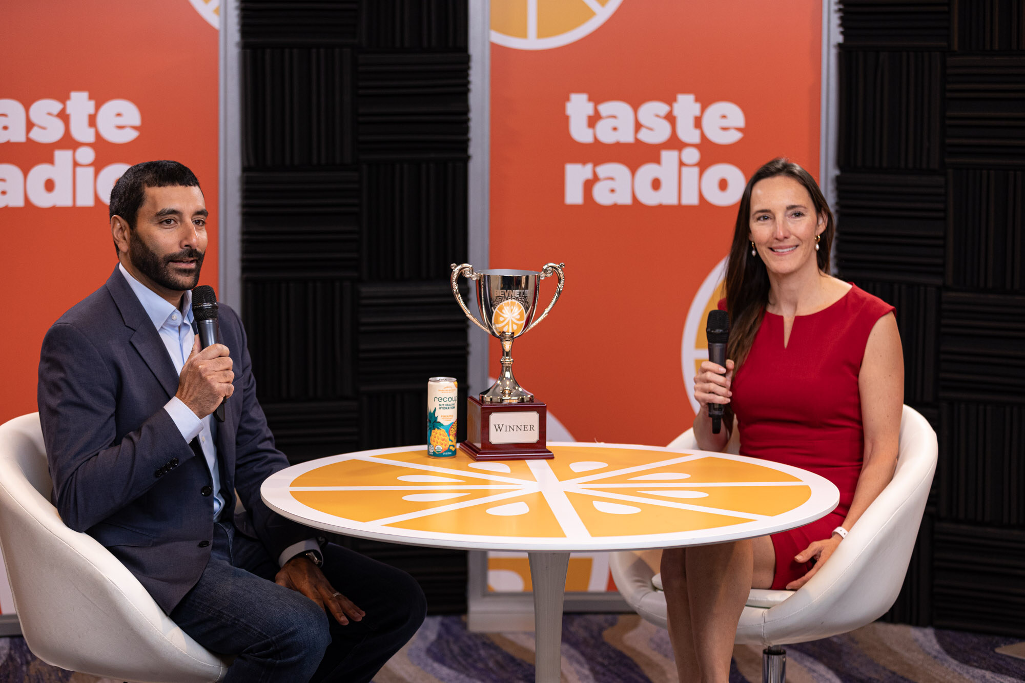 A man in a navy suit and a woman in a red dress sit at a round table with a trophy and a can in front of a Taste Radio backdrop.