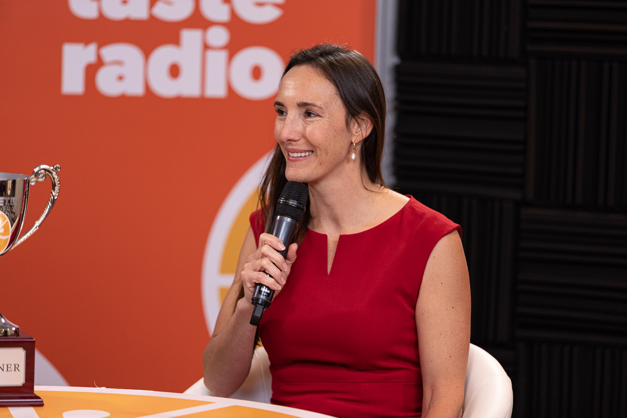 Woman in a red dress holds a microphone and smiles at a Taste Radio table with a trophy beside her.