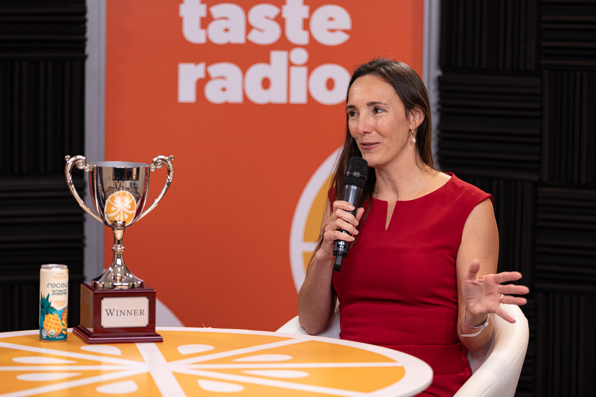 Woman in a red dress holds a microphone and gestures at a table with a Winner trophy and a can.